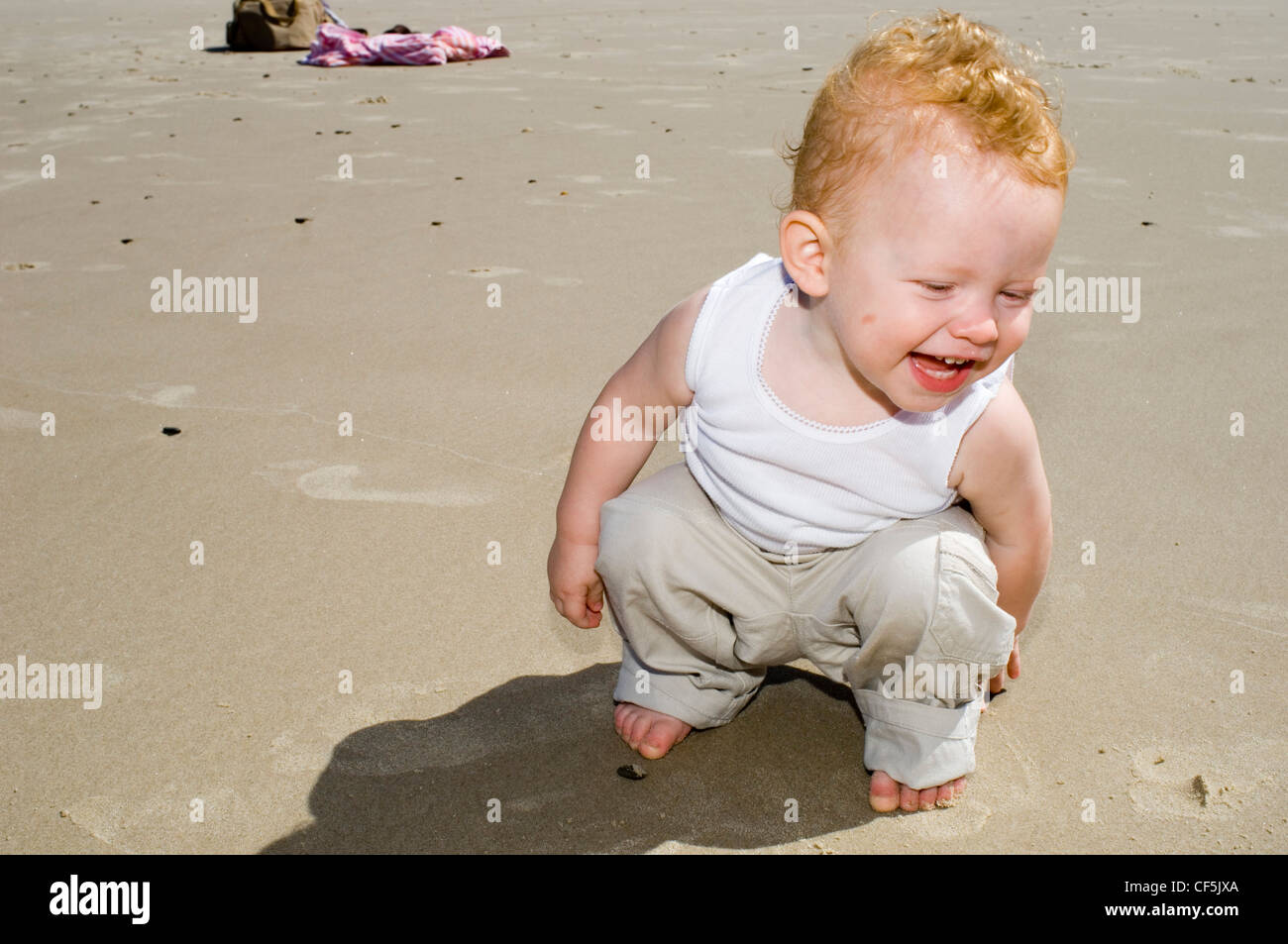 Männliches Baby mit blonden Haaren, trägt eine weiße Weste und Beige Hose,  sitzt am Strand, Lächeln Stockfotografie - Alamy