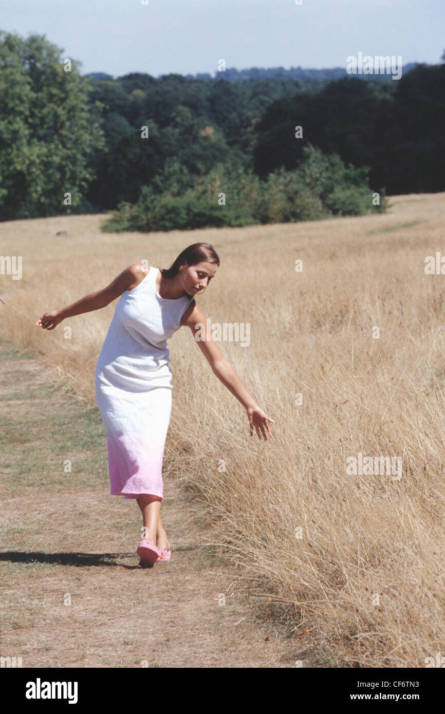 Weibliche lange brünette Haare, weiße Kleid Rosa an Unterseite und rosa Flip flops, zu Fuß in Feld übergreifen Kommissionierung Stockfoto