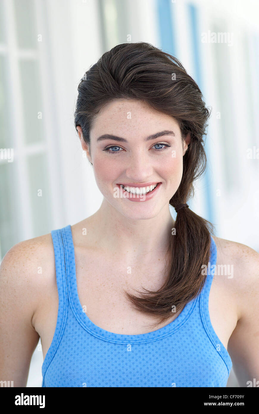 Frau mit lange brünette Haare zu einem Pferdeschwanz tragen ein blaues ärmelloses Top und machen sich in Brauntönen, lächelnd in die Kamera Stockfoto