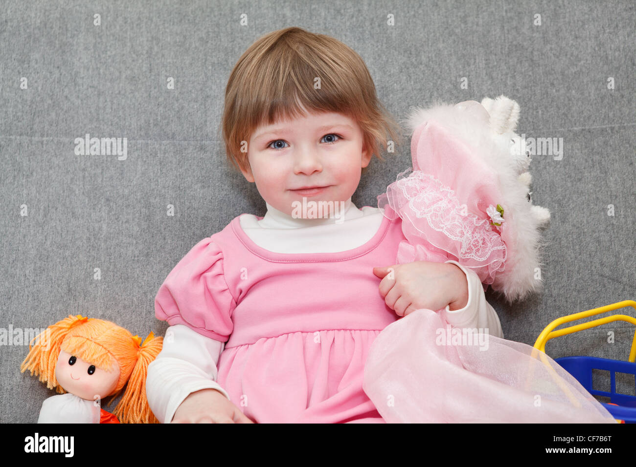 Porträt des kleinen kaukasischen russische Mädchen im rosa Kleid sitzt auf der Couch mit Puppen und lächelnd Stockfoto