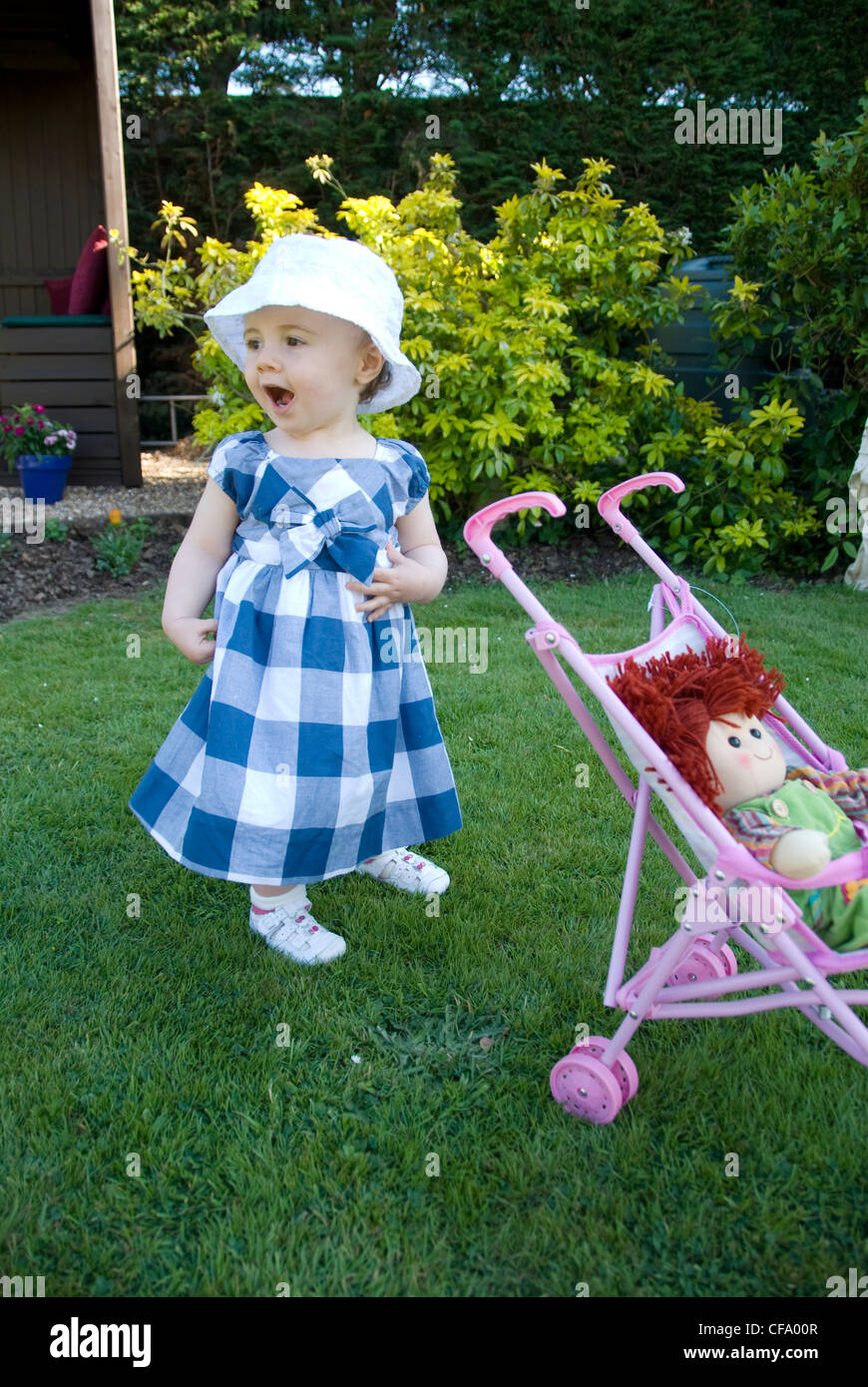 Ein weibliches Baby trägt einen weißen Sommerhut, pink, weiß Trainer und blau kariertes Kleid, stehend auf dem Rasen im Garten, Stockfoto