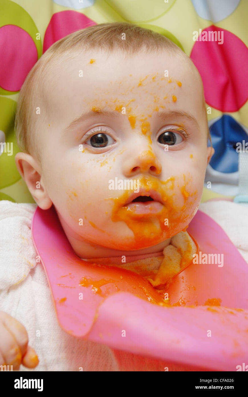 Ein weibliches Baby blondes Haar, im Hochstuhl sitzen, tragen eine quadratische Musselin und rosa Kunststoff-Lätzchen, püriert Essen Orange gefärbt Stockfoto