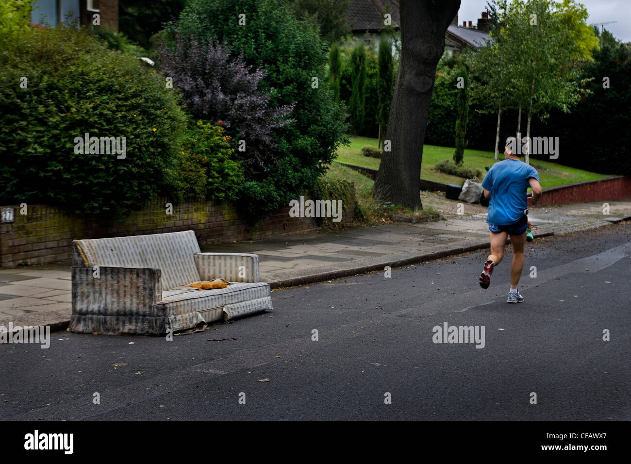 Mann in blauen Hosen und Tshirt joggt entlang einer Straße, wo ein altes Sofa auf der Straße, Highgate, London weggelassen worden Stockfoto