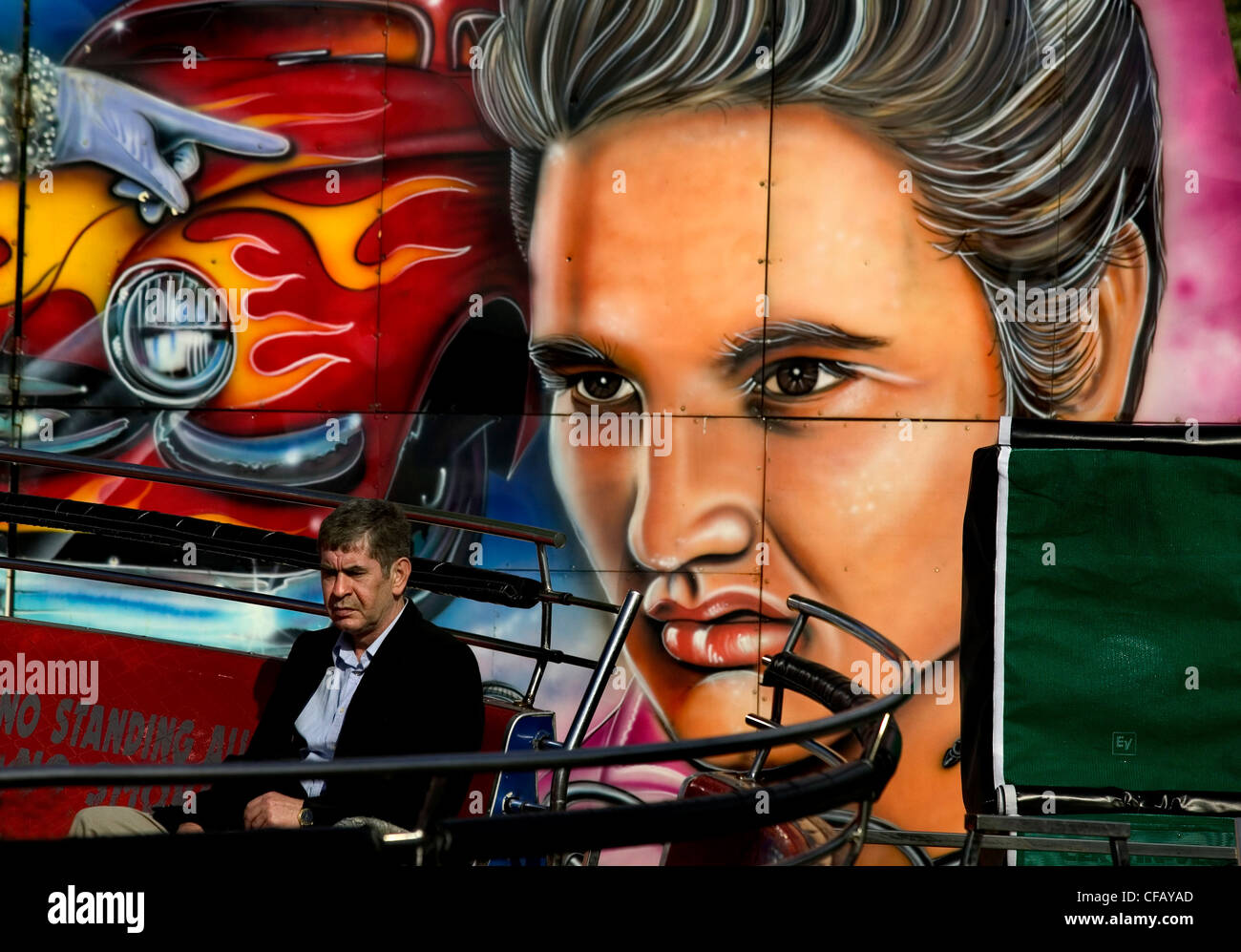 Festplatz am Hampstead Heath, London. Mann sitzt auf der Bank vor Wand Wandbild mit Gesicht von Elvis Presley und Auto Stockfoto