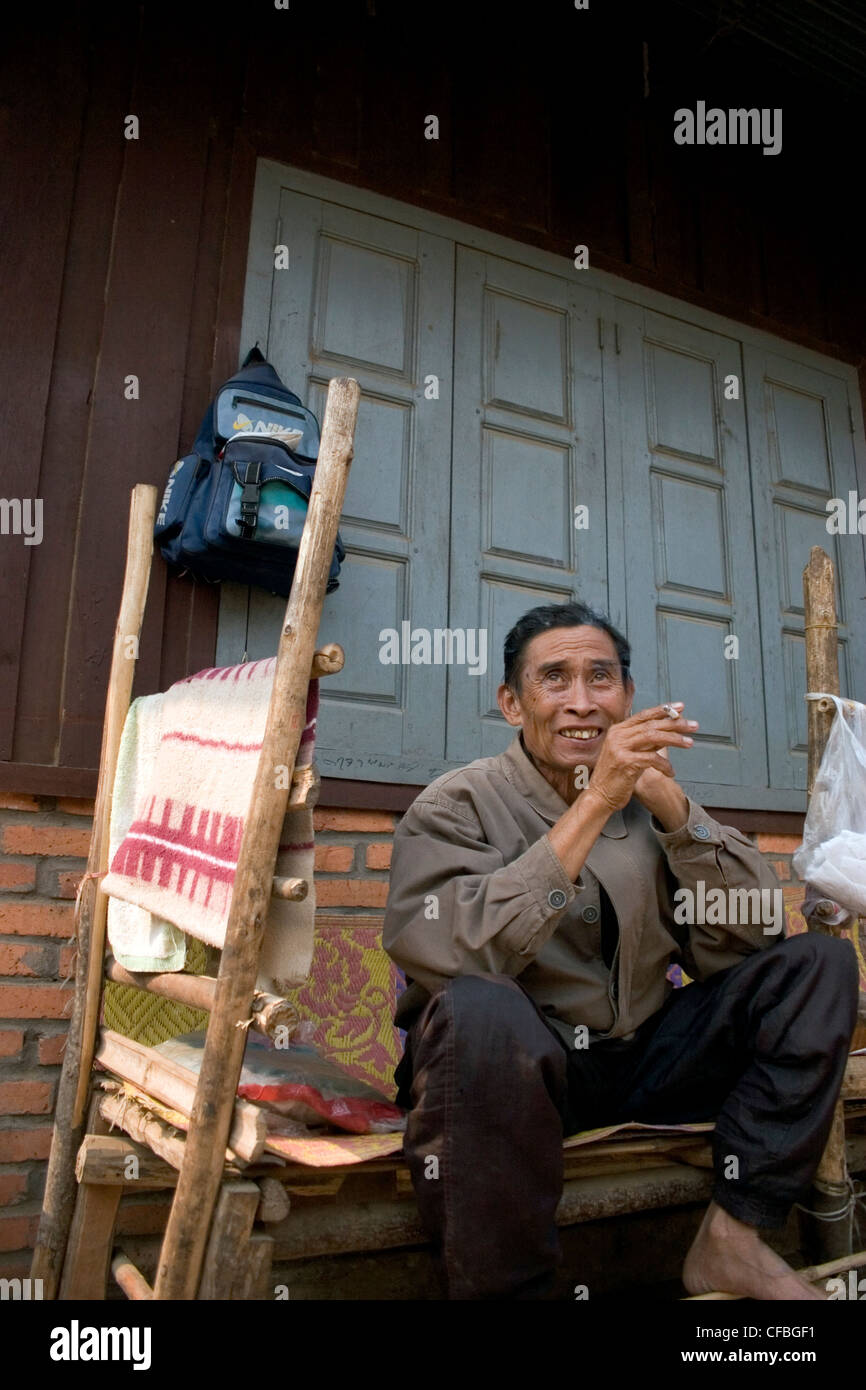 Ein asiatischer Mann mittleren Alters sitzt auf einer Bank, rauchte eine Zigarette in einem Heim in Luang Namtha, Laos. Stockfoto