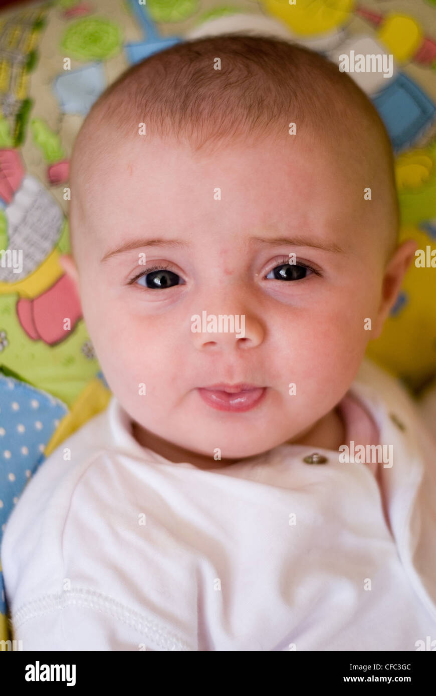 Ein weibliches Baby gekleidet in eine weiße Babygrow gegen ein Multi farbige Kissen sitzen mit ihren Lippen geschürzt Stirnrunzeln Stockfoto