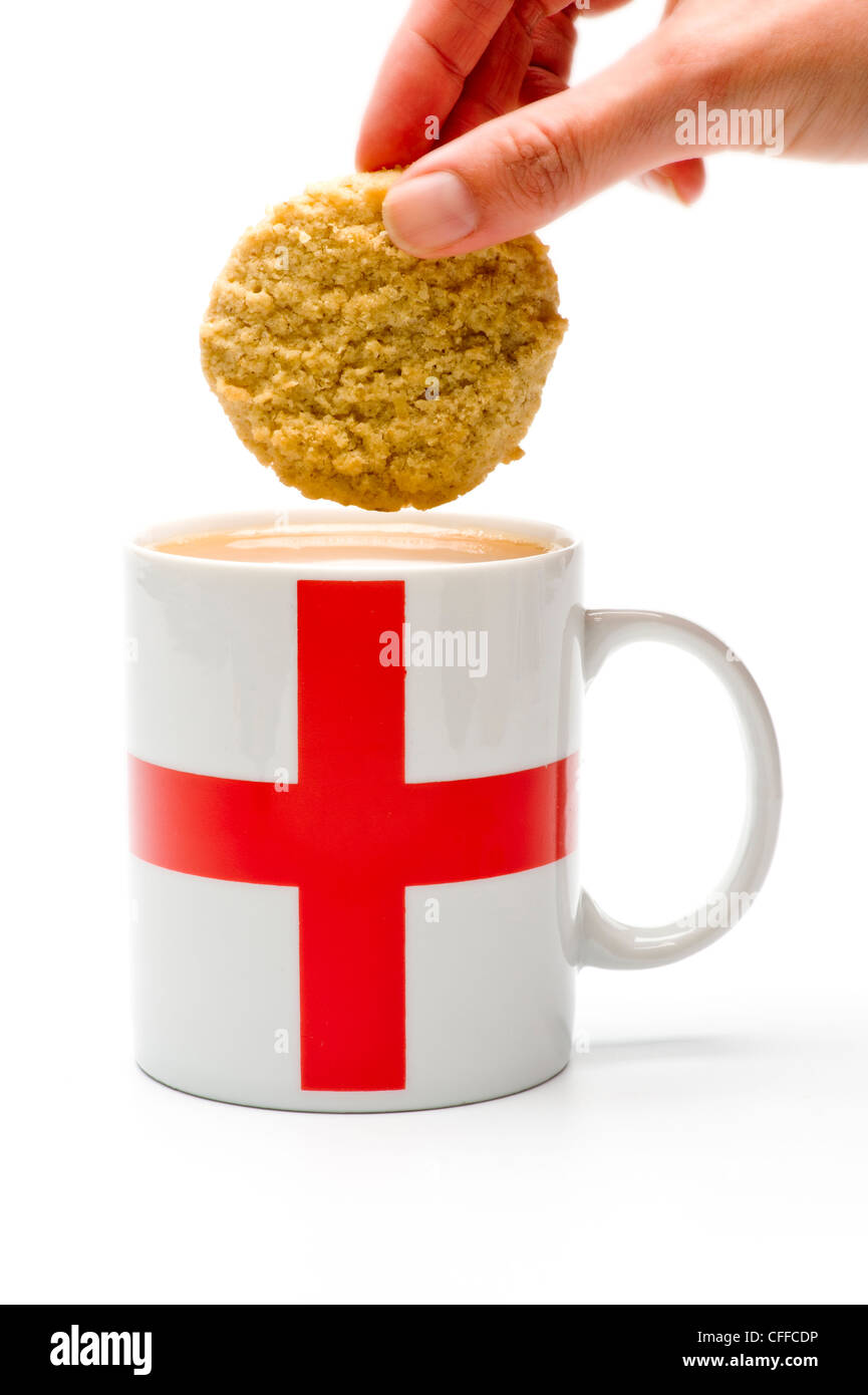 Kaukasische Hand, die einen Keks hält, der bereit ist, in einen weißen Becher mit einer englischen Flagge auf der Seite, gefüllt mit Tee, zu tauchen. Stockfoto