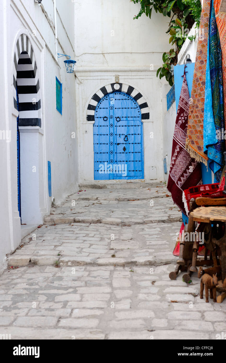 Sidi Bou Said. Tunesien. Blick auf Souvenir-Teppiche zum Verkauf an schmale gepflasterte Gasse mit schwarzen und weißen gewölbten blaue Tür an Stockfoto