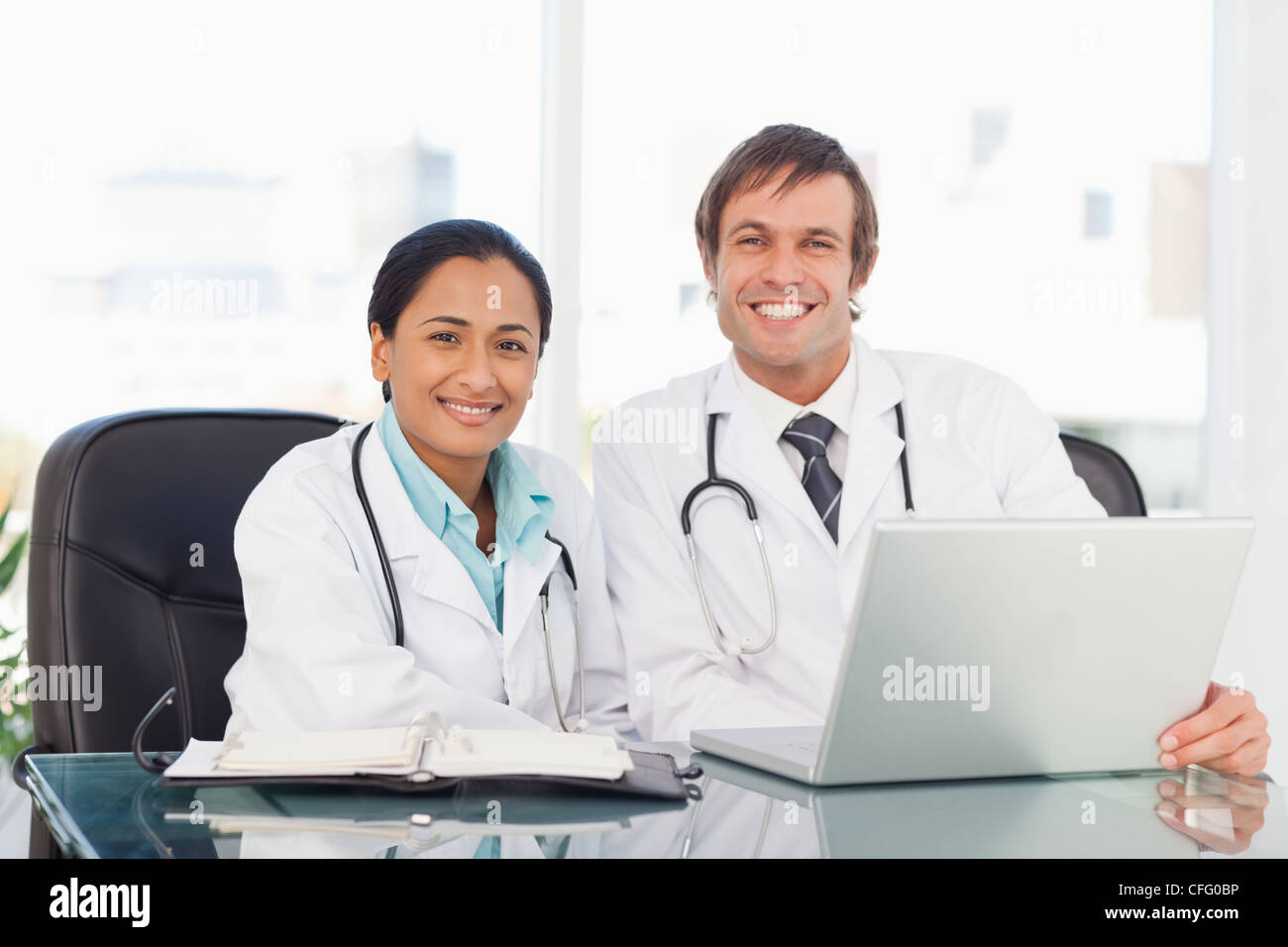 Lächelnde Arzt sitzend vor seinem Laptop und wird begleitet von einem Kollegen Stockfoto