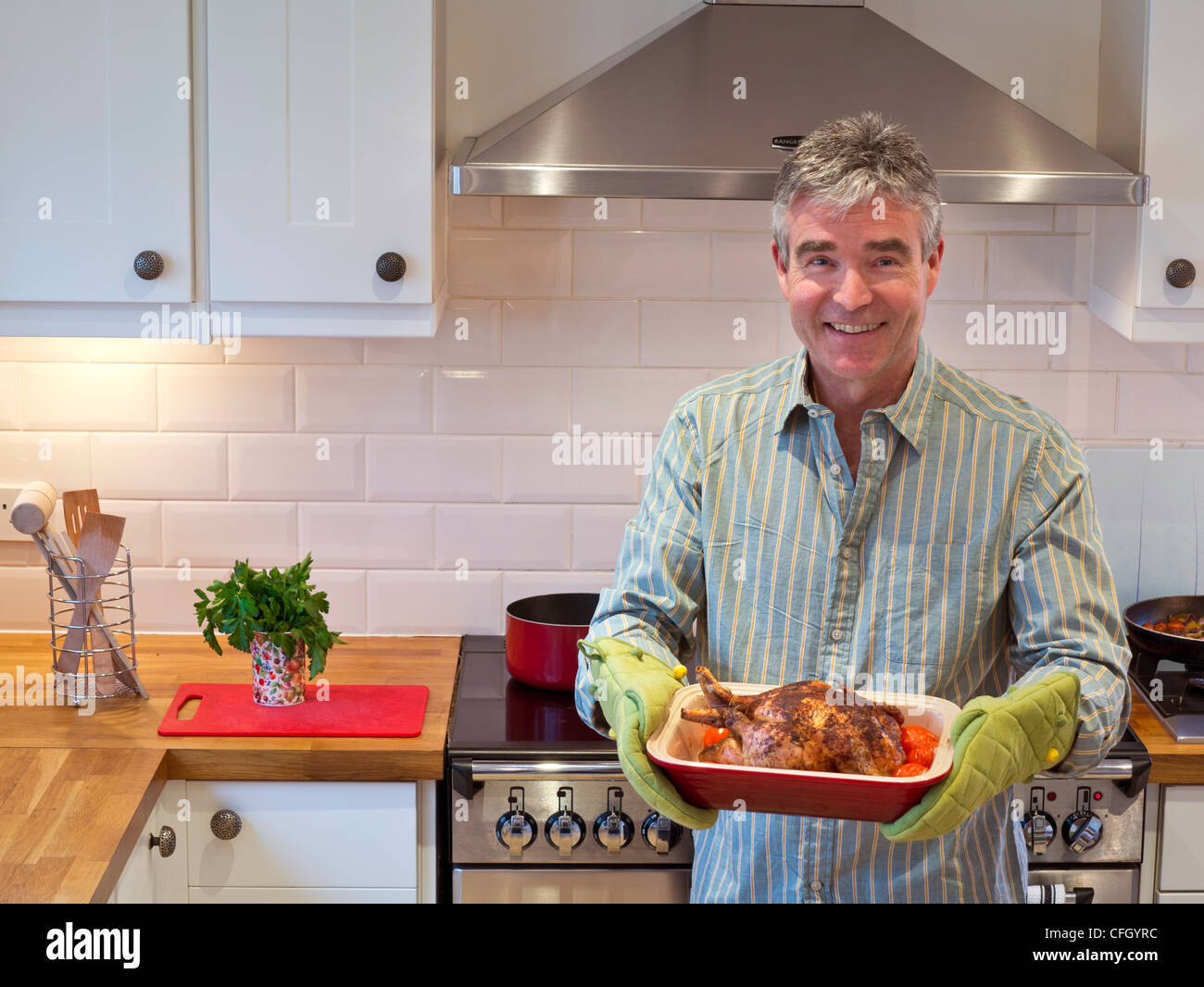 Mann KOCHEN HUHN Lächeln Entspannt zuversichtlich reifer Mann in der zeitgenössischen Küche bietet eine warme, frisch gekochte Mahlzeit von Hähnchen und Gemüse Stockfoto