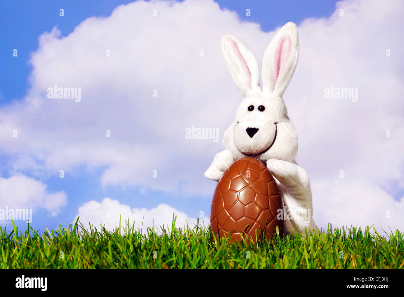 Spaß Foto einer weißen Ostern Hase Marionette hält ein große Schokoladen-Ei auf dem Rasen mit einem blauen bewölkten Himmelshintergrund. Stockfoto