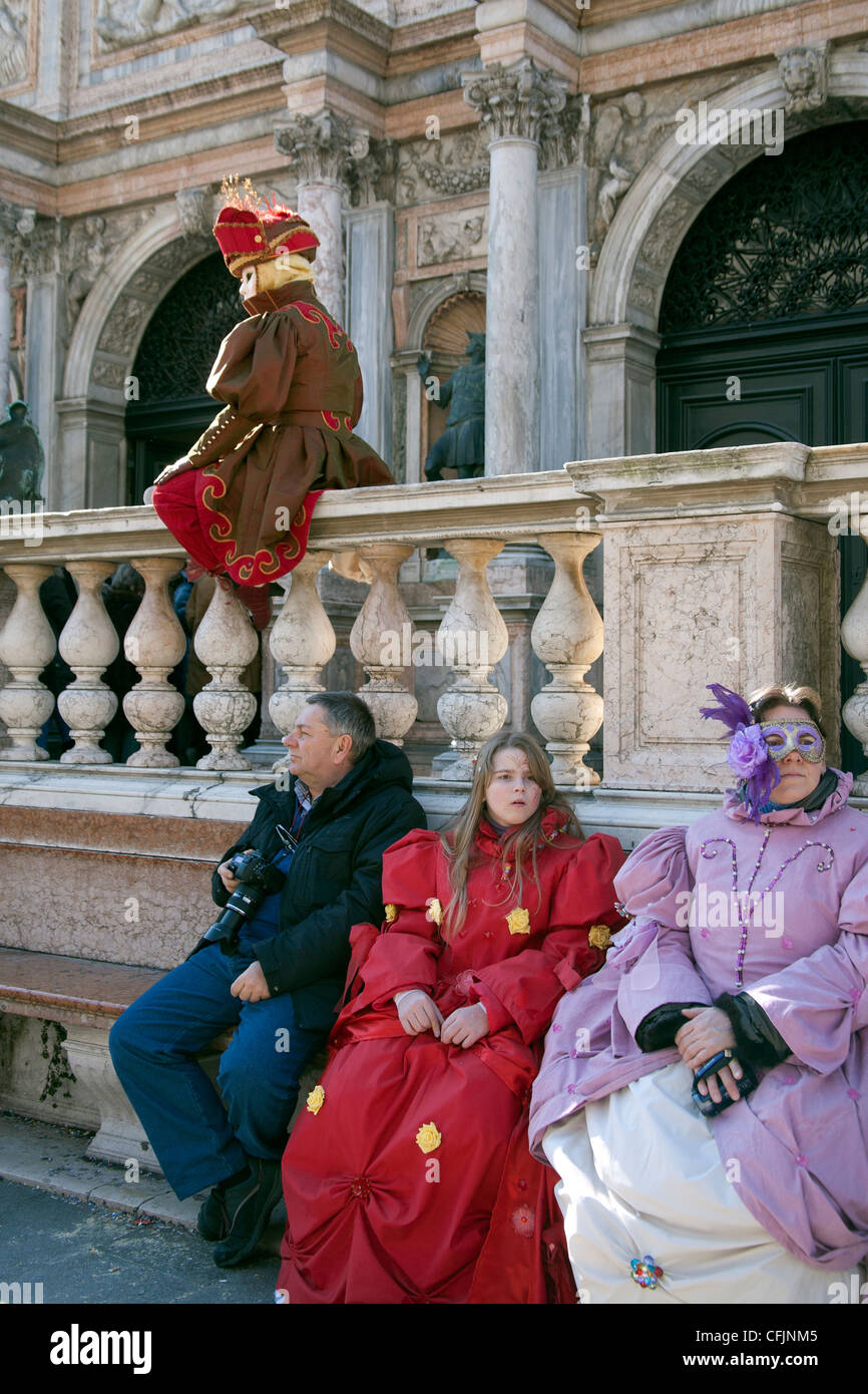 Familie im Karnevalskostüm sitzen auf einer Bank schauen gelangweilt als Karneval Charakter Posen auf einer Wand in Ihrer Nähe St. Marks Platz Venedig Stockfoto