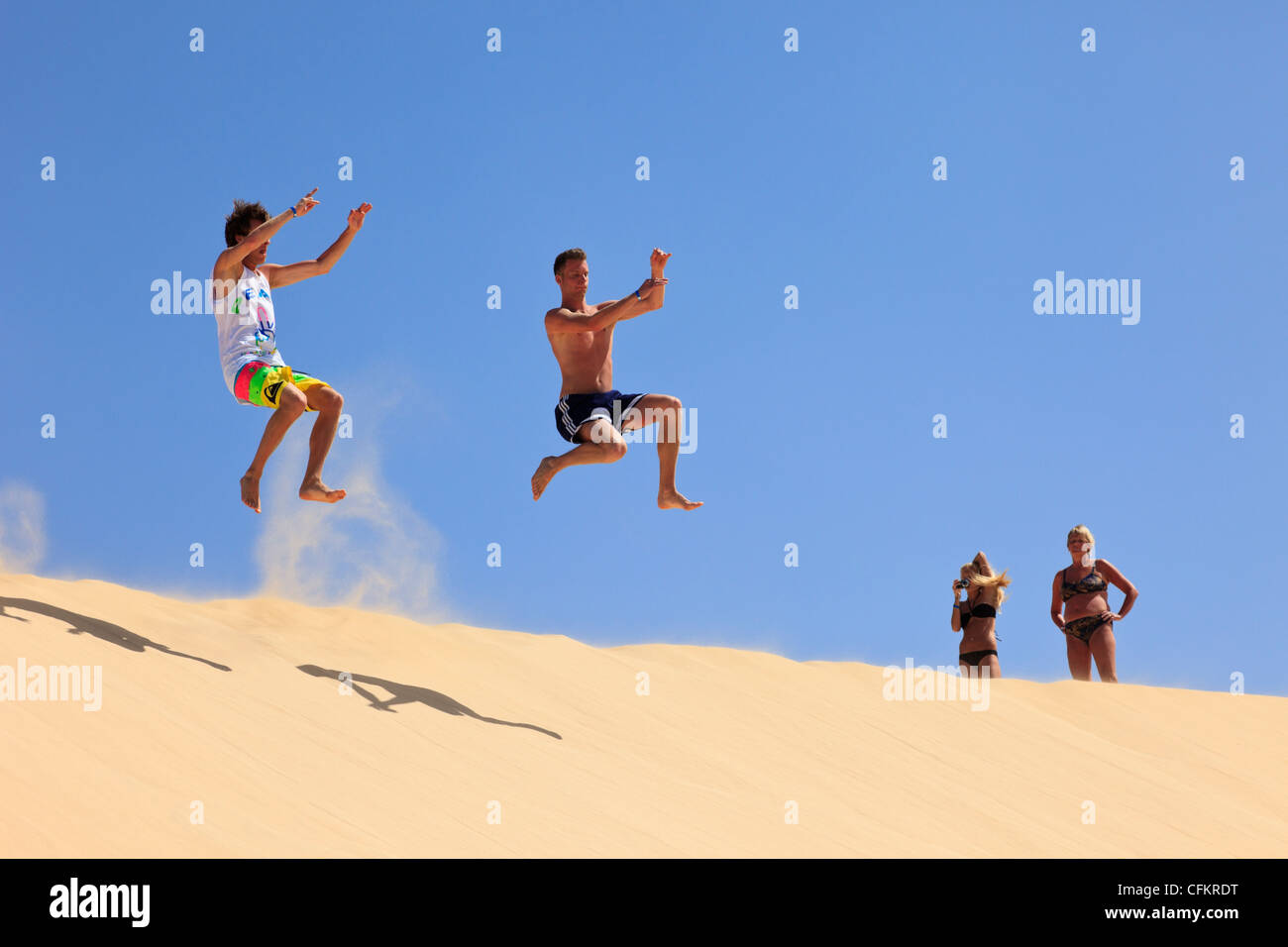 Zwei Männer springen in der Luft von einer großen Sanddüne mit zwei Frauen beobachten und fotografieren an einem sonnigen Tag. Boa Vista, Kapverdische Inseln Stockfoto