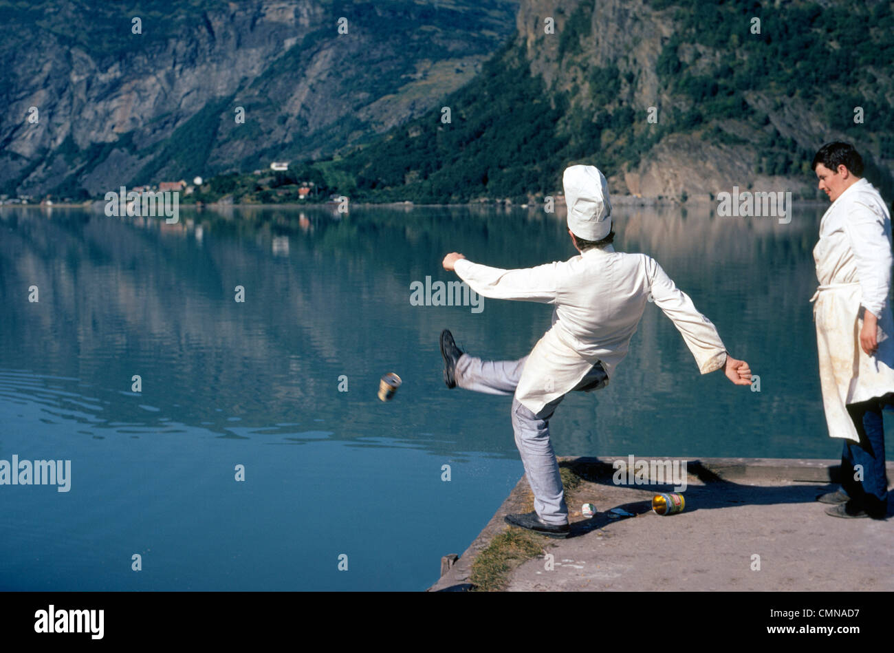 Zwei männliche Küchenchefs verschmutzen einen schönen skandinavischen Fjord durch Tritte Blechdosen eins zu einem Zeitpunkt in das malerische Gewässer in der Nähe von Elveseter, Norwegen. Stockfoto