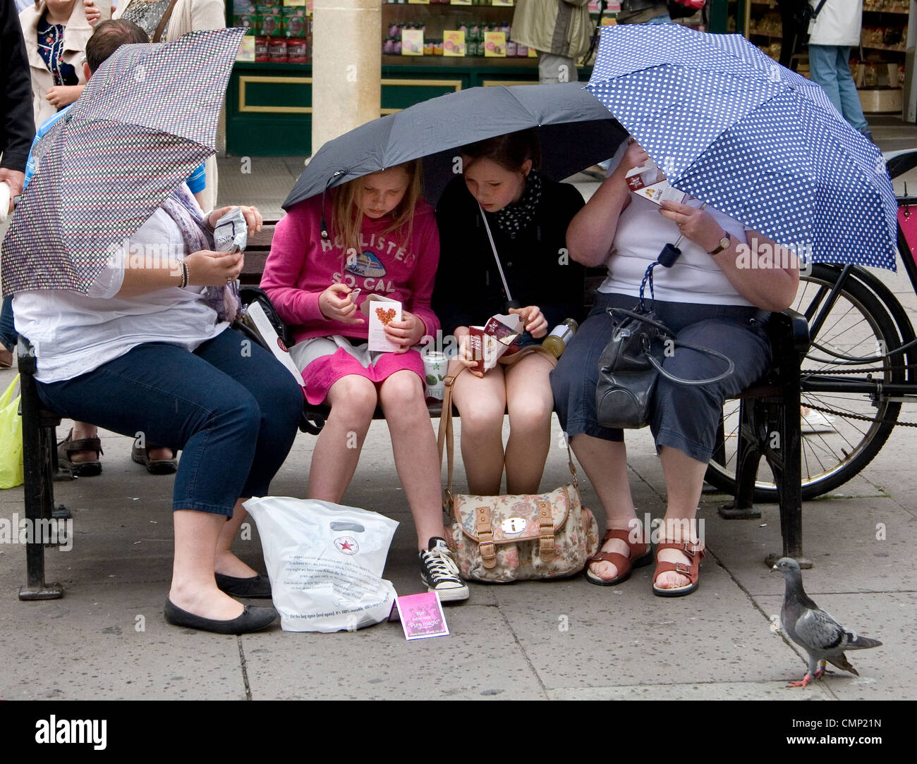 2 Frauen mit 2 Kindern Essen Mittagessen auf einer Straße Bank während bergende unter Sonnenschirmen; eine Taube Uhren. Stockfoto