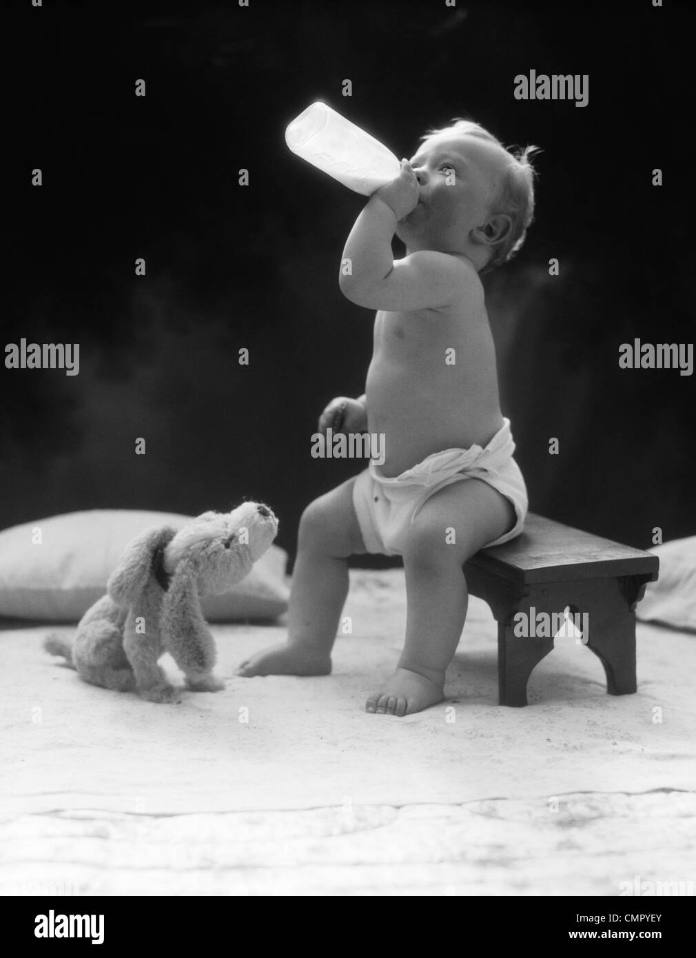 1930ER JAHRE BABY SITZEND AUF STUHL TRINKEN MILCH AUS DER FLASCHE MIT AUSGESTOPFTEN HUND BLICKTE ZU IHM Stockfoto