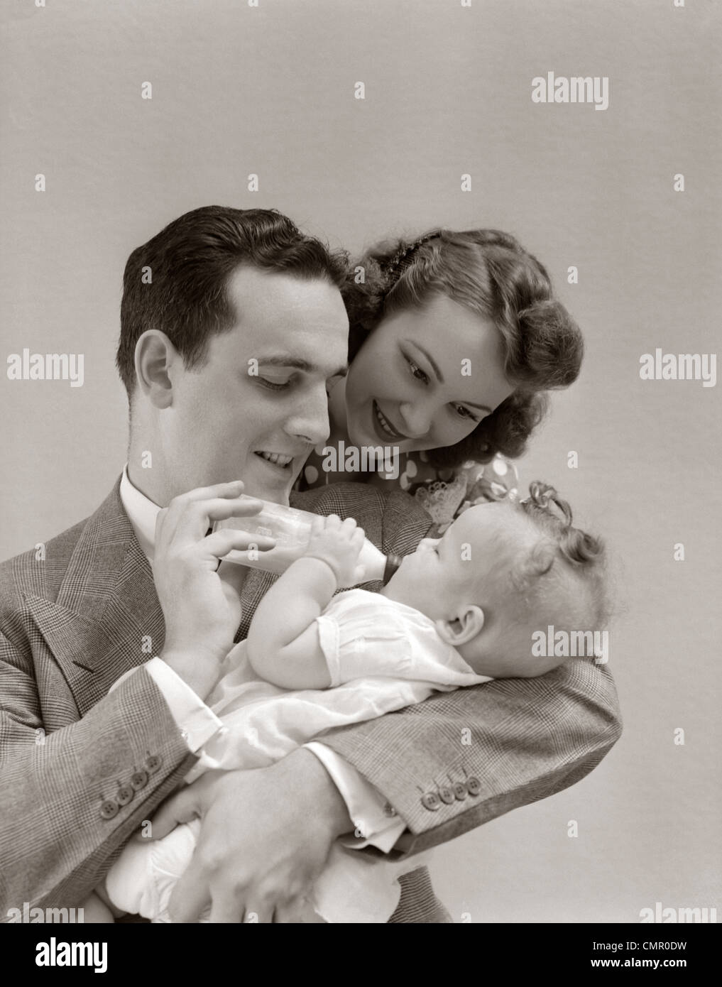 1940ER JAHREN VATER WIEGT BABY TOCHTER FÜTTERN IHRE FLASCHE MIT MUTTER ÜBER SEINE SCHULTER BLICKEND Stockfoto