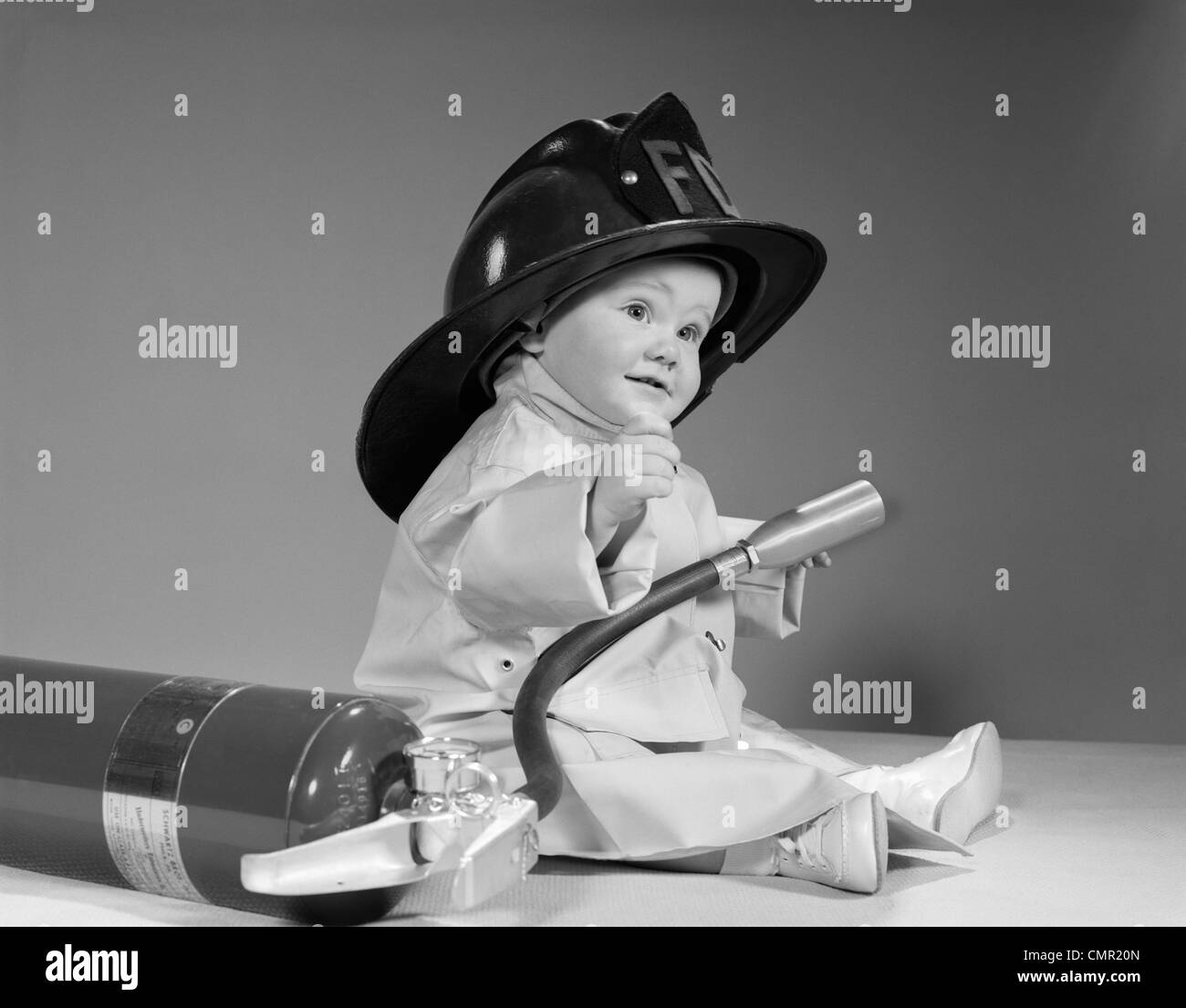 1960ER JAHRE BABY FEUERWEHRMANN MIT SICHERHEIT HUT MANTEL UND FEUERLÖSCHER Stockfoto