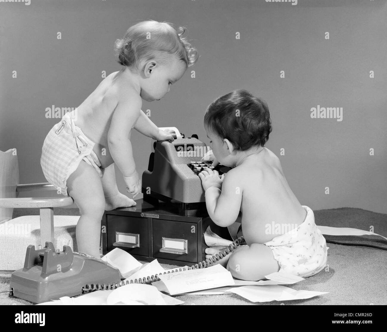 1960ER JAHREN PAAR BABYS UM RECHENMASCHINE MIT TELEFON OFF HOOK & PAPIERE AUF BODEN VERSTREUT Stockfoto