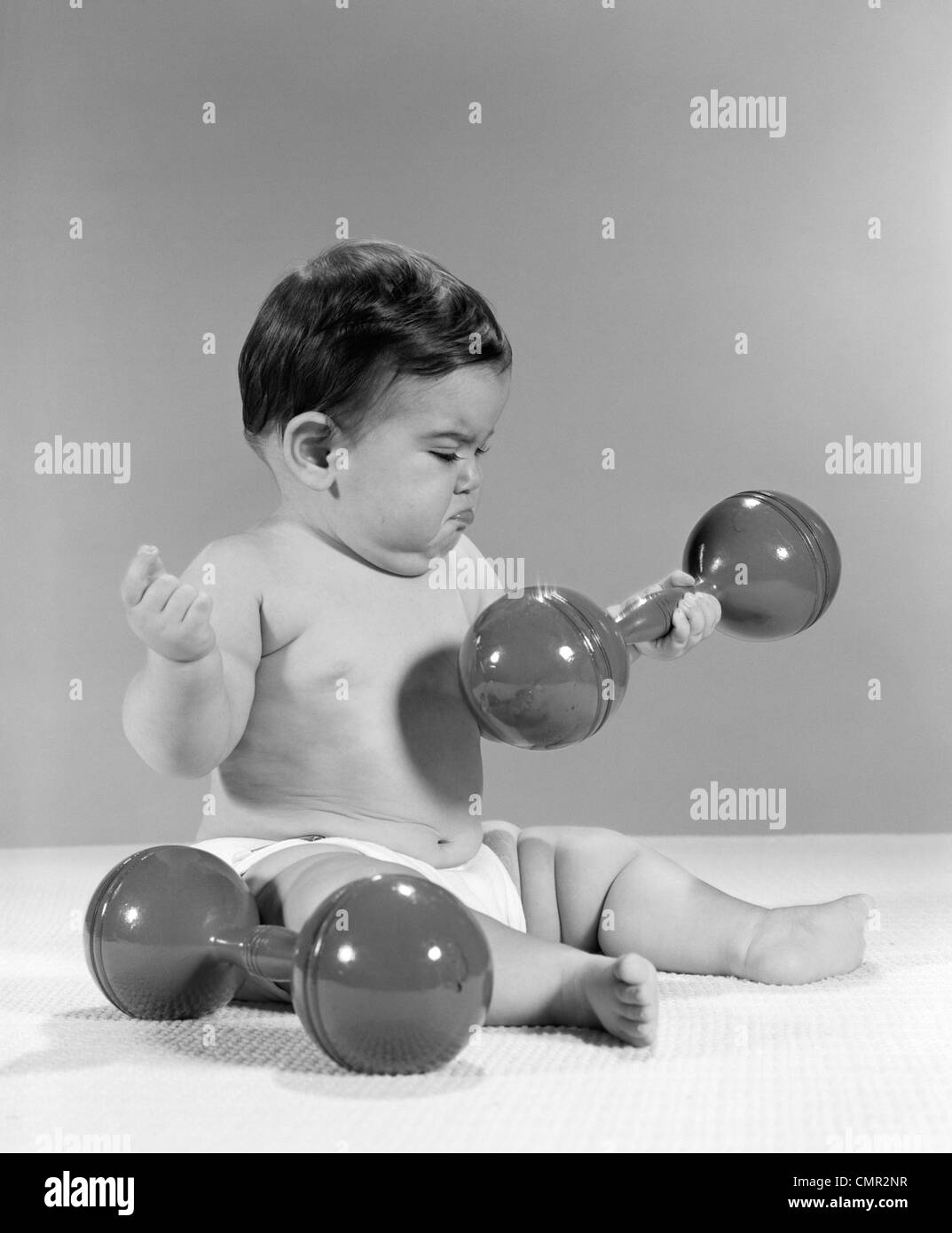 1960ER JAHRE BABY SITZEND HEBEN EINE HANTEL MIT EINEM ANDEREN SEITLICH MIT MÜRRISCHEN AUSDRUCK IM GESICHT Stockfoto