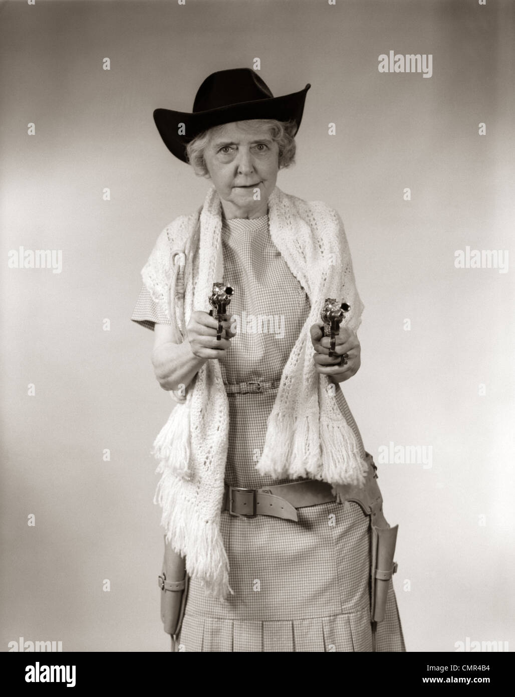 1950ER JAHRE OMA COWGIRL HUT & SCHAL TRAGEN & ZEIGT 2 PISTOLEN UND BLICK IN DIE KAMERA Stockfoto