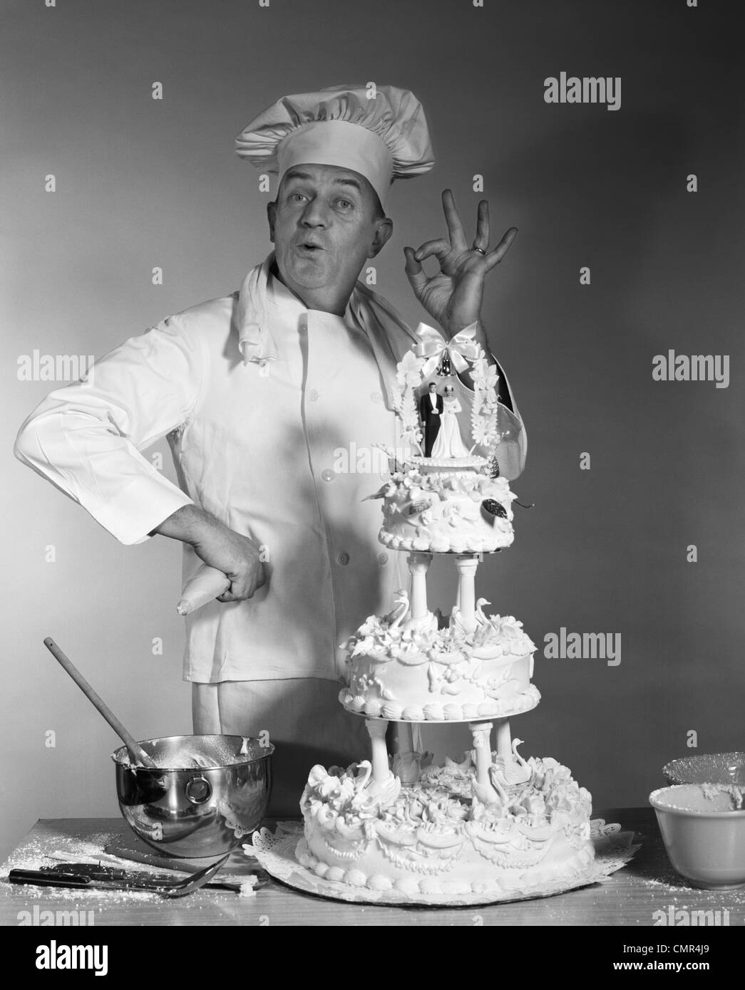 1950ER 1960ER JAHRE 1970ER JAHREN MANN PORTRÄT BAKER BLICK AUF KAMERA MACHEN OK HANDZEICHEN NEBEN DREI TIER HOCHZEITSTORTE BRAUT & BRÄUTIGAM ON TOP Stockfoto