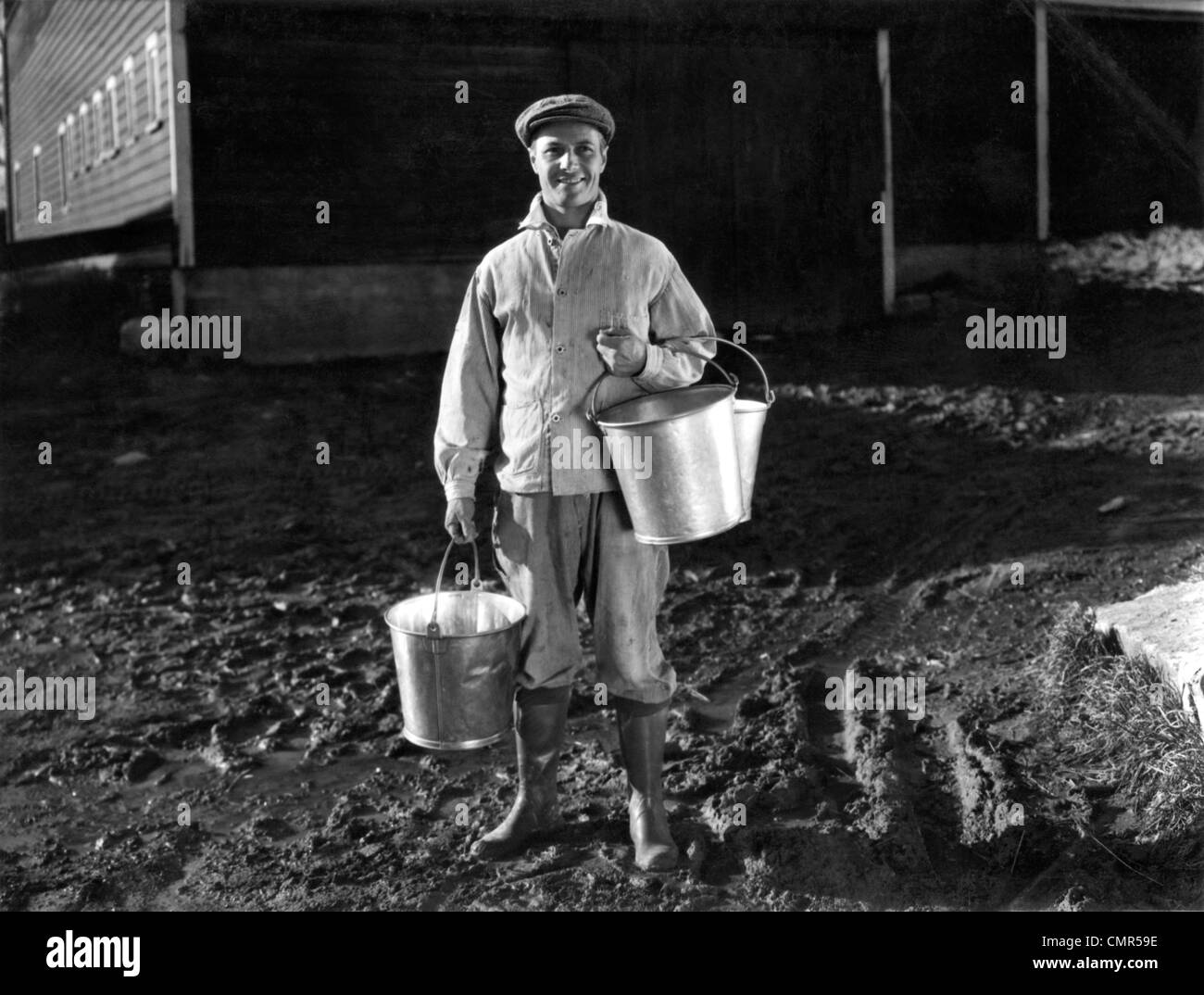 1930S 1940S STIEFEL MANN BAUER SENNER AUßERHALB SCHEUNE HÄLT MILCH EIMER TRAGEN MÜTZE JACKE RUBBER KNIE LÄCHELNDEN BLICK IN DIE KAMERA Stockfoto