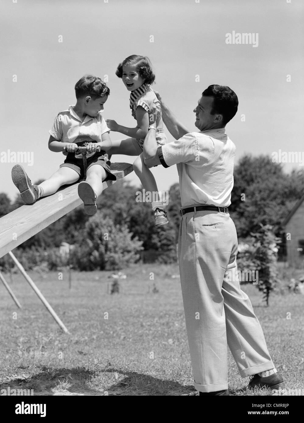 1950ER JAHREN VATER HEBEN SOHN UND TOCHTER AUF EINEM SPIELPLATZ WIPPE IM FREIEN Stockfoto