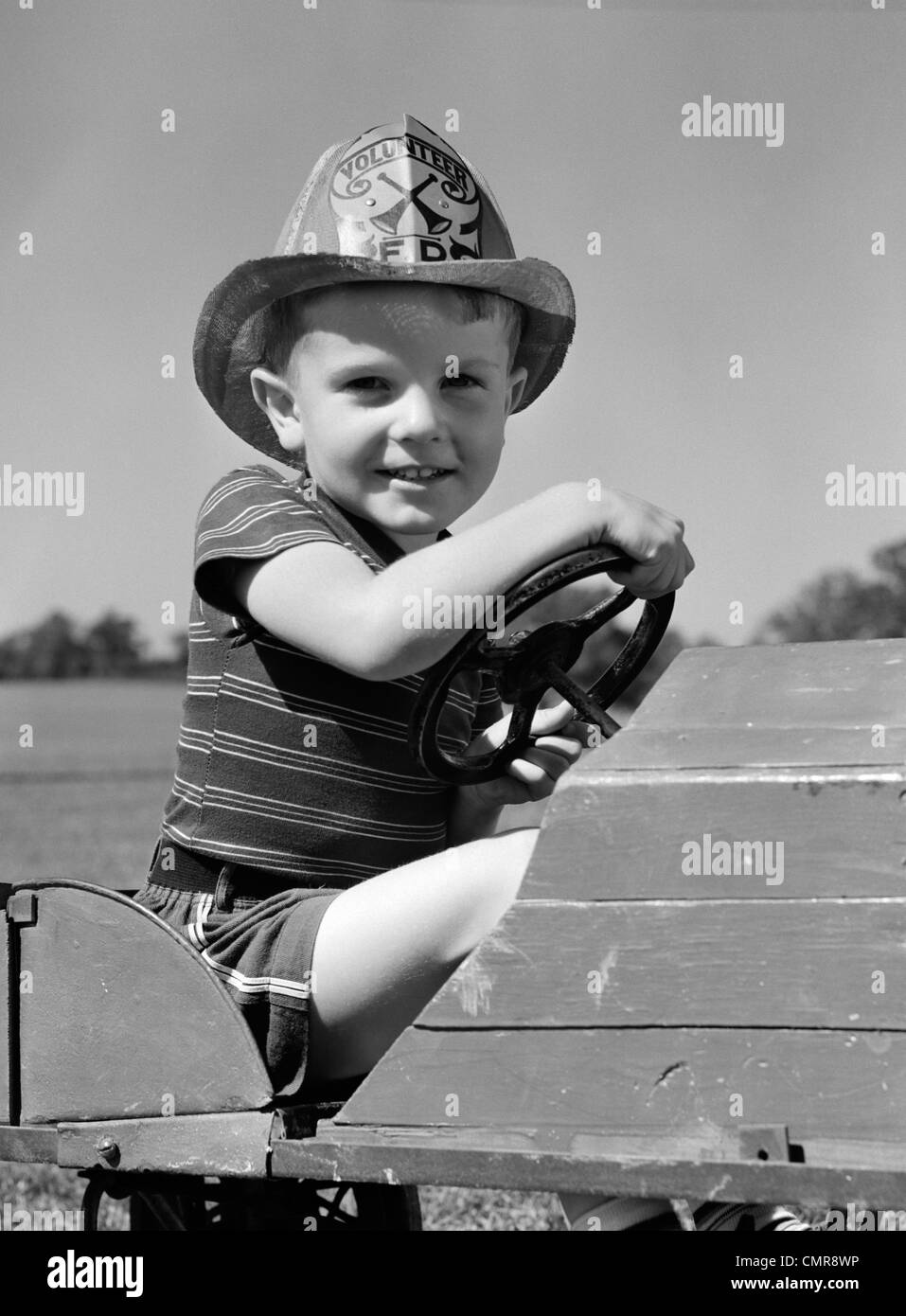 1940ER JAHRE JUNGEN SPIELEN FEUERWEHRMANN IN SPIELZEUG FEUER LKW TRAGEN FIREMAN ES SICHERHEIT HUT HÄLT LENKRAD LÄCHELND Stockfoto