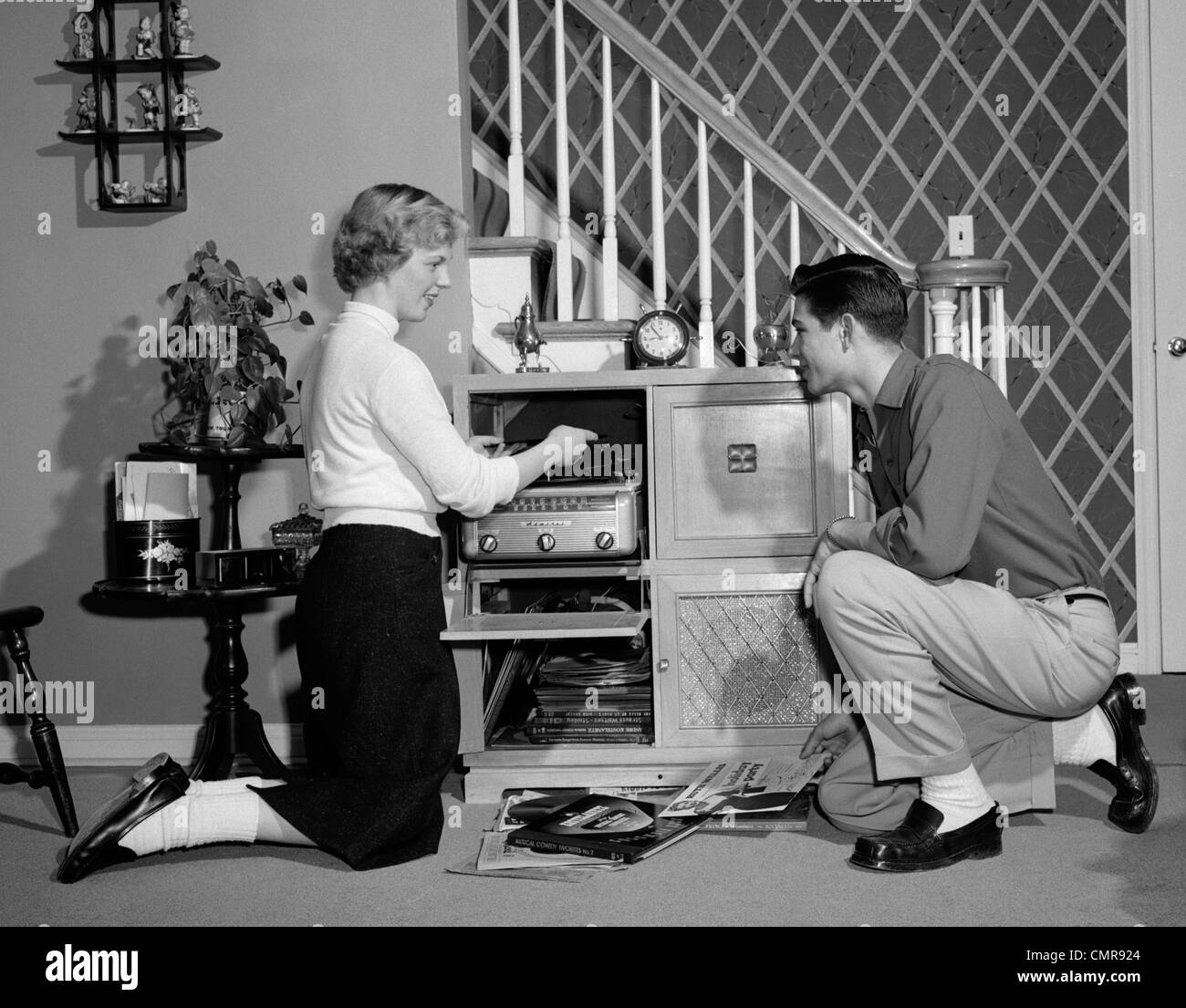 1950ER JAHRE TEENAGER-PAAR SPIELEN HI-FI-DATENSÄTZE AUF KONSOLE PHONOGRAPHEN IM WOHNZIMMER Stockfoto
