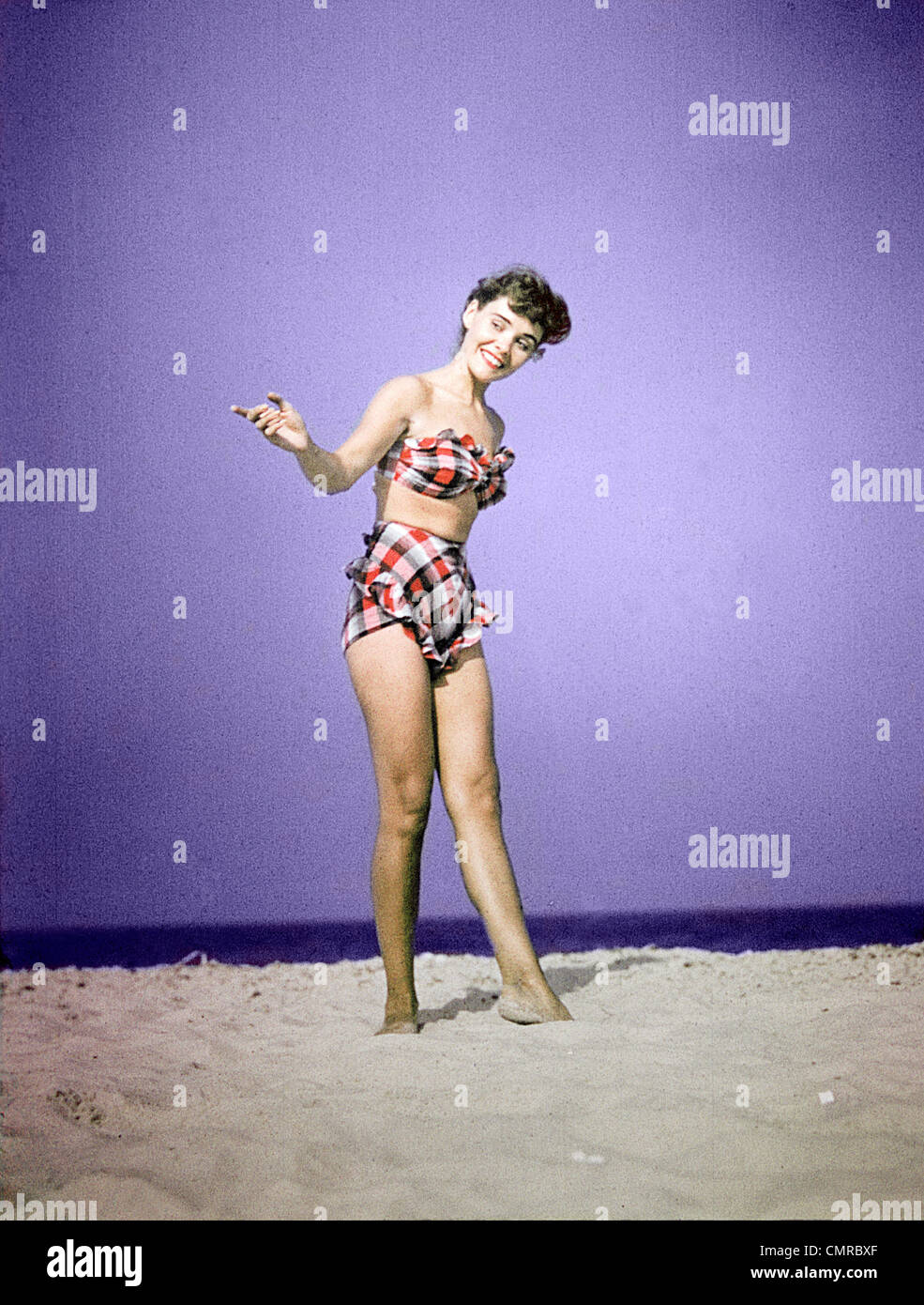 1940S 1950S LÄCHELNDE JUNGE FRAU TRAGEN PLAID ZWEI STÜCK BADEANZUG AM STRANDSAND Stockfoto