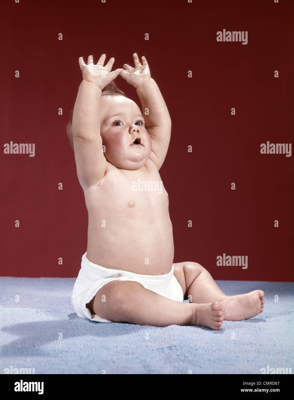 1960ER JAHREN HOBEN BABY TRAGEN TUCH WINDEL SITZEN DIE ARME ÜBER DEN KOPF Stockfoto