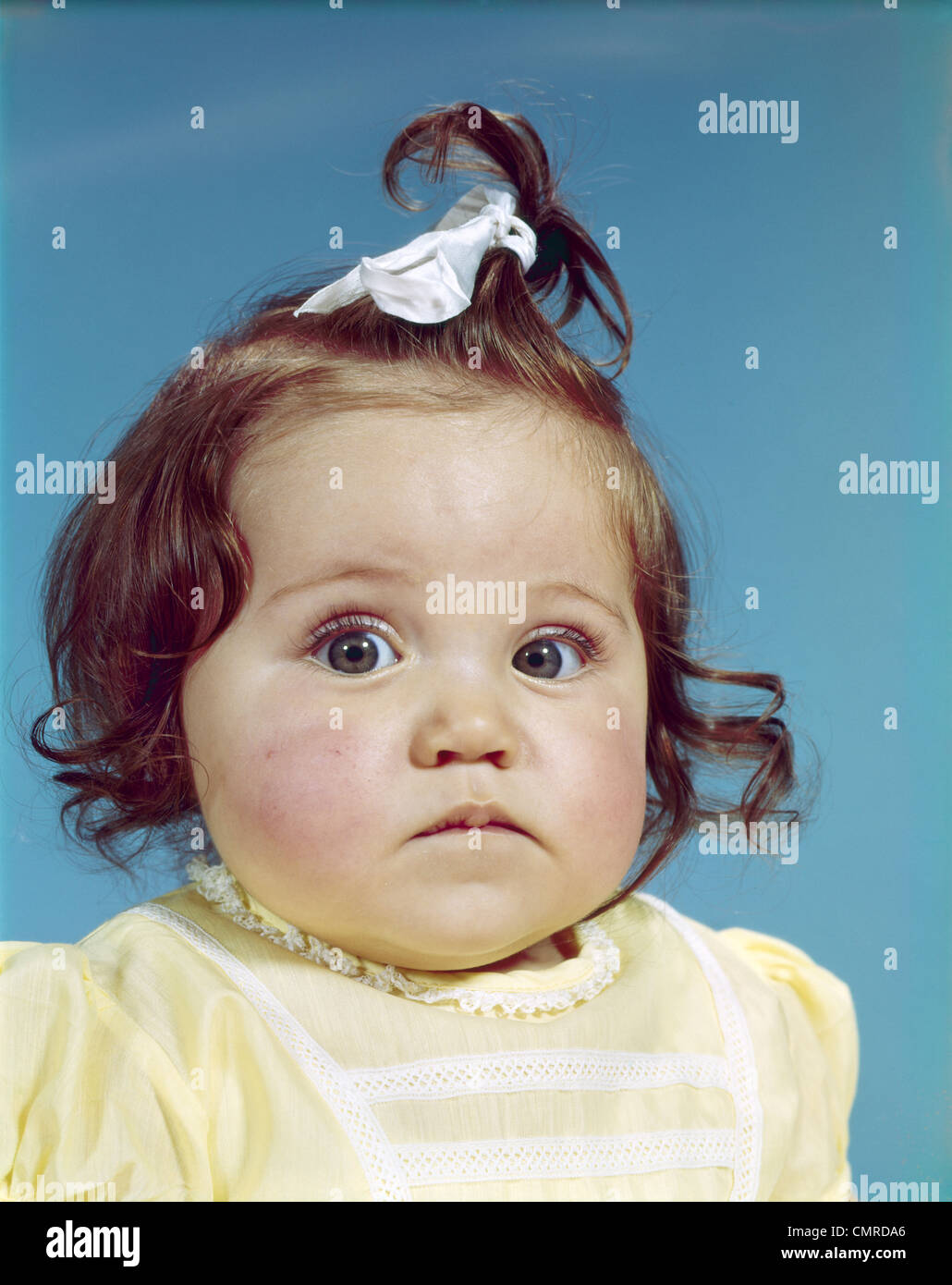 1960ER JAHRE BRÜNETTE BABY GIRL BAND HAARKNOTEN GELBE SHIRT MOLLIG WANGEN RUNDES GESICHT TRAURIG UNGLÜCKLICH GESICHTSAUSDRUCK BLICK IN DIE KAMERA Stockfoto