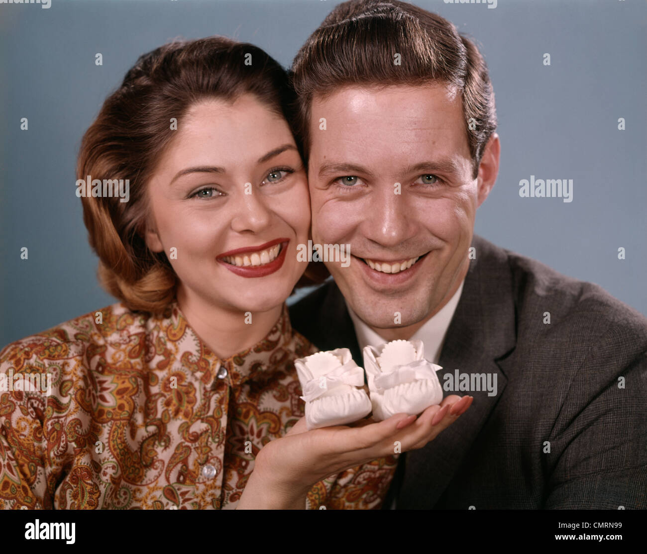 LÄCHELNDE PAAR HALTEN BABY SCHUHE FAMILIE VATER MUTTER STUDIO DER 1960ER JAHRE Stockfoto