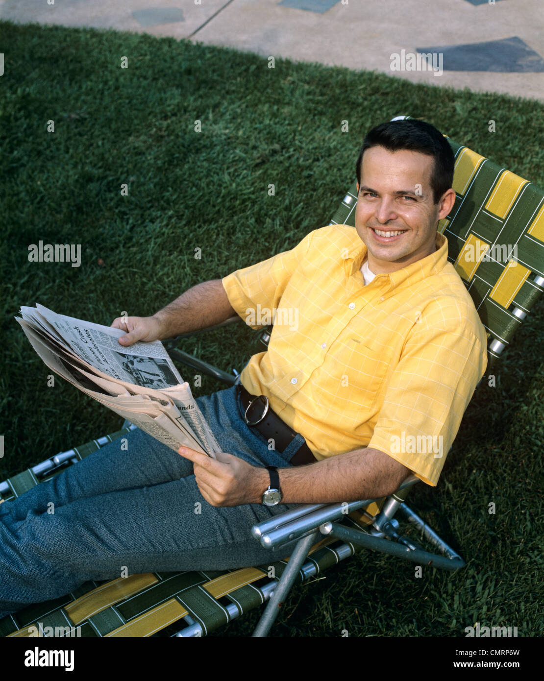 1970 1970S RETRO-LÄCHELND MANN SITZT IM RASEN STUHL LESEN ZEITUNG ENTSPANNENDE FREIZEIT IM FREIEN Stockfoto