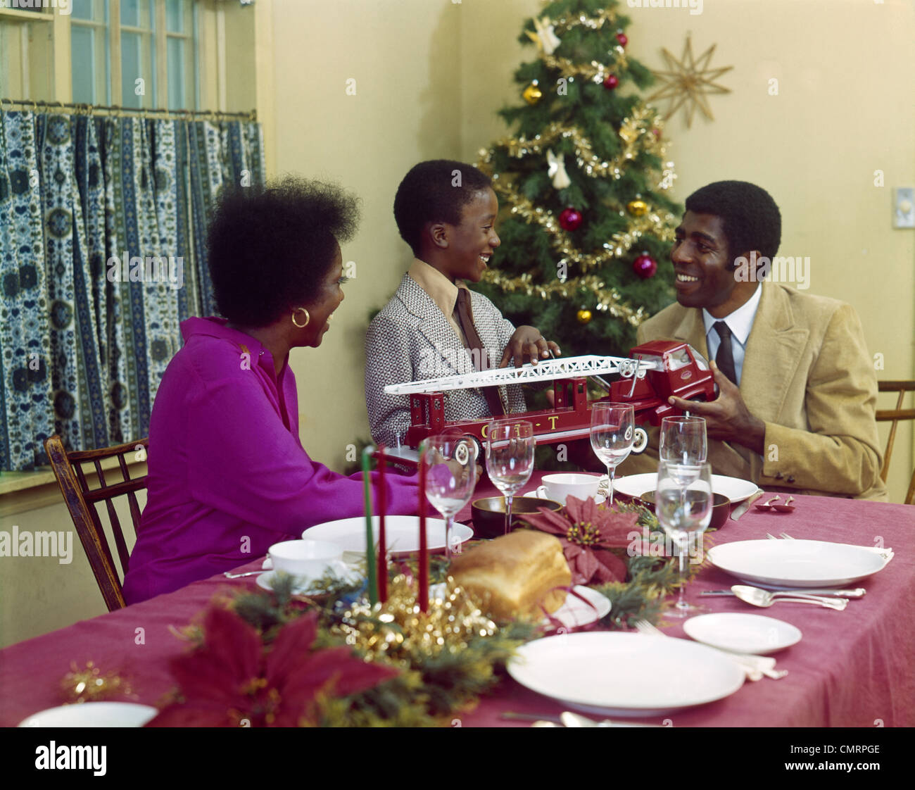 1970ER JAHRE AFRO-AMERIKANISCHE FAMILIE MUTTER VATER SOHN ESSTISCH MIT SPIELZEUG-FEUER-LKW-WEIHNACHTSBAUM Stockfoto