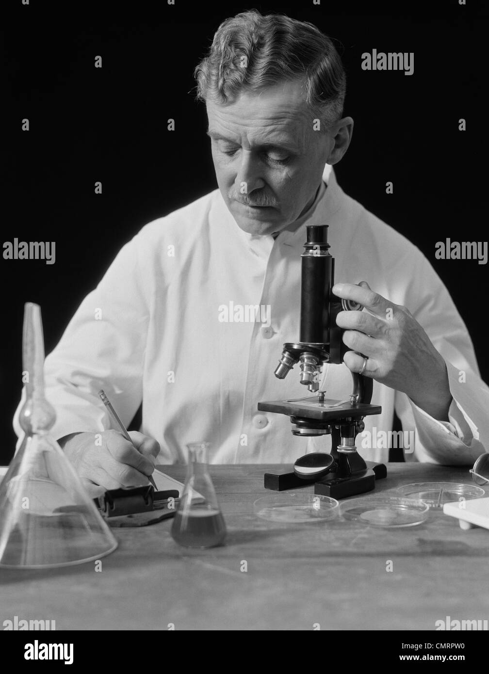 1920S 1930S MÄNNLICHE WISSENSCHAFTLER TRAGEN WEIßE LAB COAT SCHREIBEN VON DATEN AUF CLIP BOARD HALTEN FOKUSSTEUERUNG DES MIKROSKOPS Stockfoto