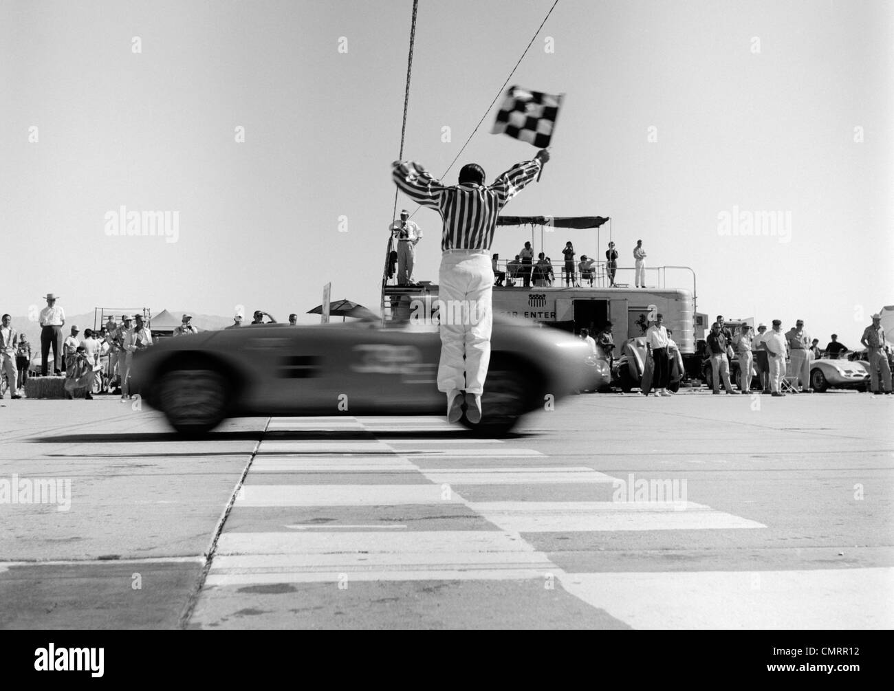 1960ER JAHRE MANN SPRINGEN WINKEN CHECKERED FLAG AS SPORTWAGEN ÜBER DIE ZIELLINIE ZU GEWINNEN Stockfoto