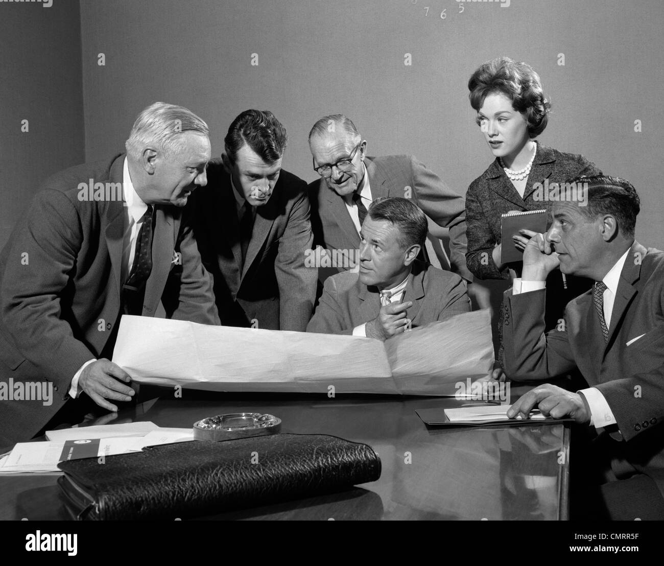 1960ER JAHRE GRUPPE 5 FÜHRUNGSKRÄFTE VERSAMMELTEN SICH UM TABELLE MIT SEKRETÄR NOTIZEN & PLÄNE VERTEILT ZWISCHEN IHNEN Stockfoto