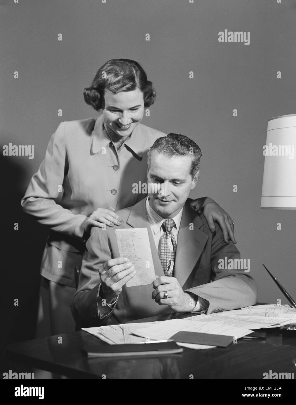 1950ER JAHREN PAAR MANN SITZT AM SCHREIBTISCH HÄLT PAPIER FRAU STEHEND MIT BLICK AUF DIE SCHULTER, LÄCHELT GLÜCKLICH Stockfoto