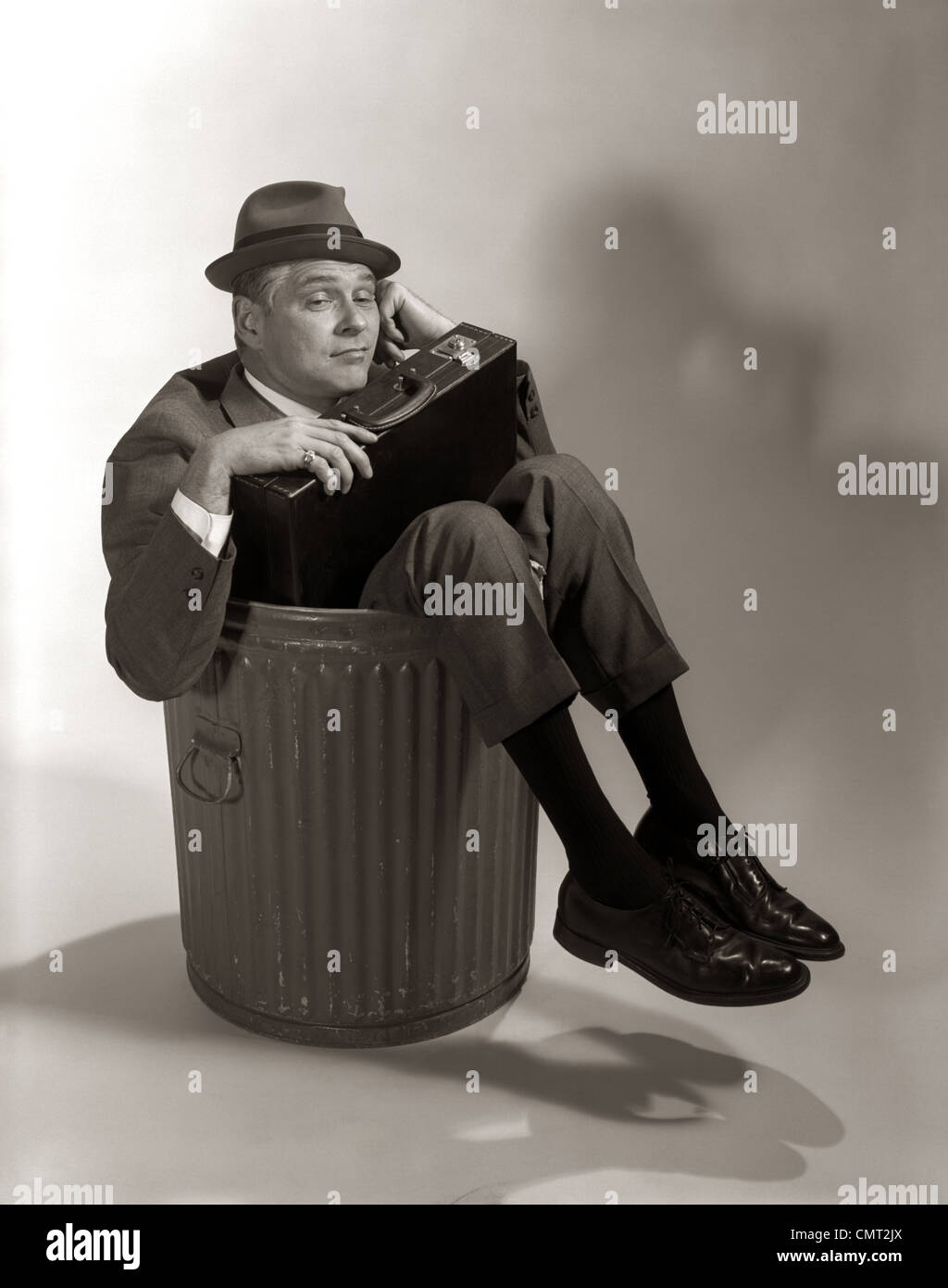 1960ER JAHREN SYMBOLISCHE GESCHÄFTSMANN IM ANZUG & HUT HÄLT AKTENTASCHE GEDUMPTEN GEBRANNTEN ENTLASSEN ENTLASSEN ARBEITSLOSEN SITZT IM MÜLLEIMER Stockfoto