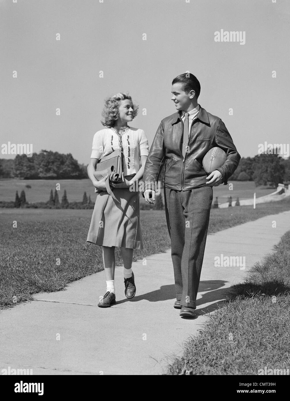 1930S 1940S TEEN PAAR HINUNTER BÜRGERSTEIG MÄDCHEN MIT SCHULJUNGE BÜCHER HALTEN FUßBALL Stockfoto