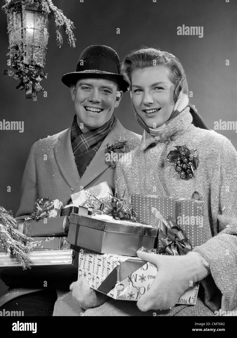 1950ER JAHREN PAAR MANN UND FRAU TRAGEN ARMLOAD DER WEIHNACHTSPAKETE LÄCHELND STEHEN IM SCHNEE UNTER DER VERANDA IM FREIEN LICHT Stockfoto