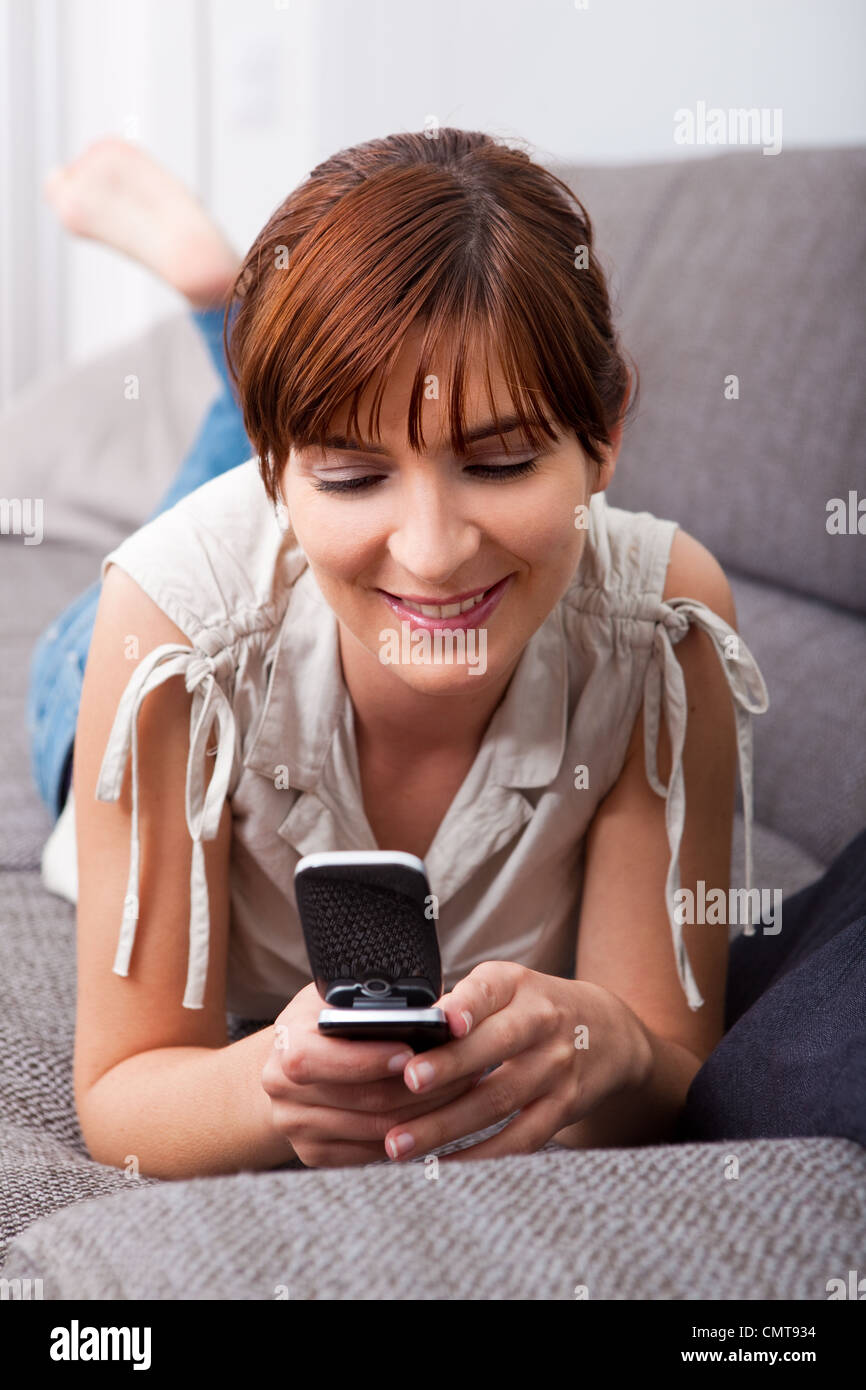 Porträt einer schönen Frau auf Sofa hält ein Handy und ein Sms-Versand Stockfoto