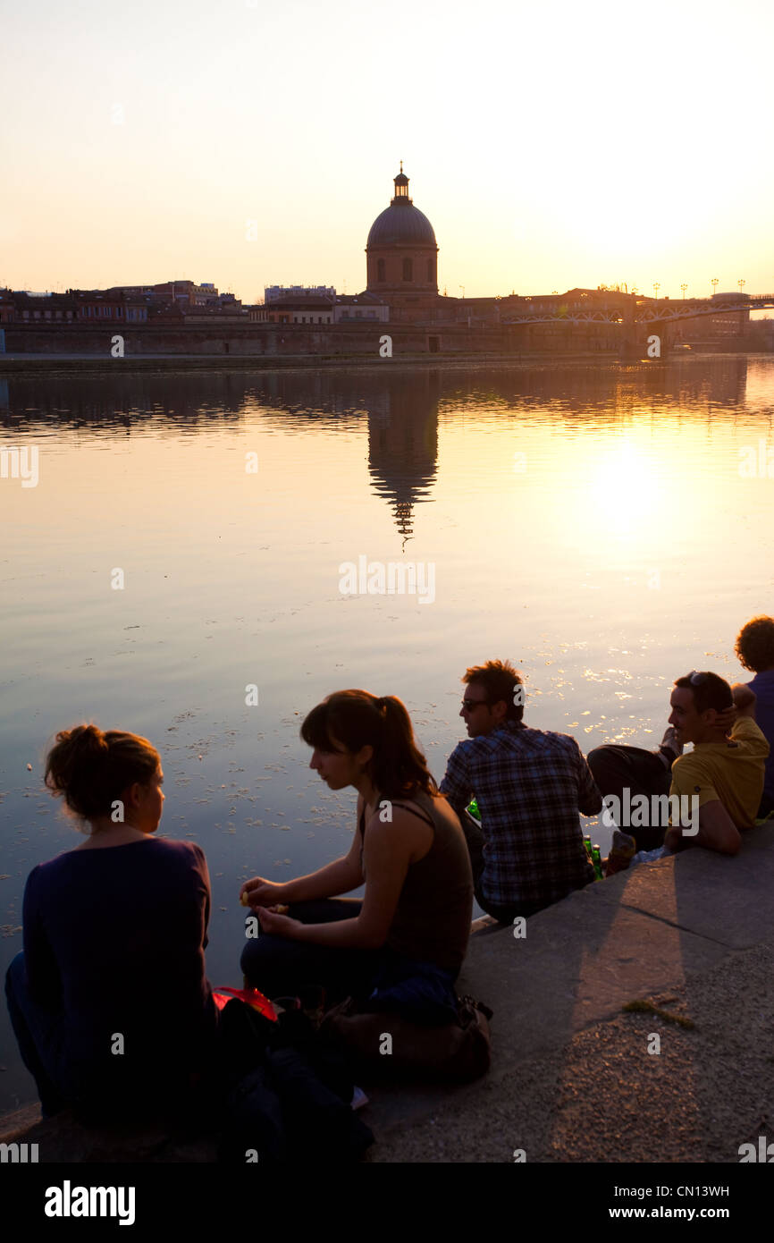 Leute sitzen am Ufer des La Garonne, wenn die Sonne dicht hinter der Kuppel De La Grave, Toulouse, Frankreich untergeht Stockfoto