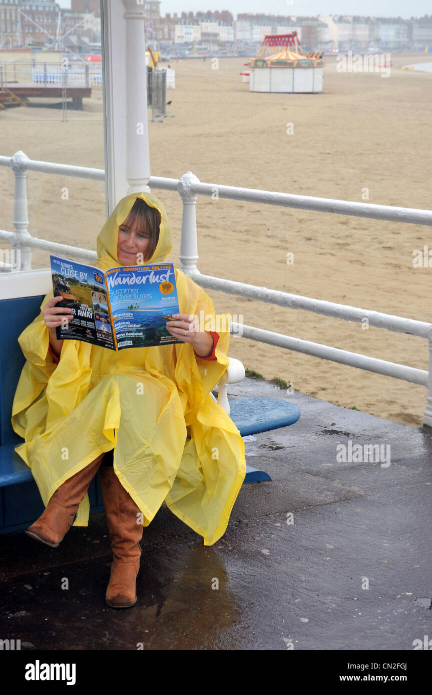 Nasses Wetter, Frau liest ein Urlaubsmagazin an einem nasskalten Weymouth Strand in Dorset, England, UK Stockfoto