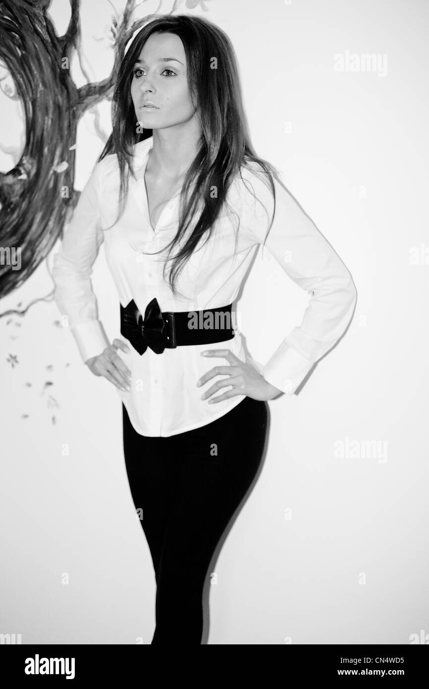 Ein einfarbiges Porträt einer besinnlichen Frau in einer stilvollen weißen Bluse mit schwarzem Gürtel, die vor der künstlerischen Kulisse eines bemalten Baumes steht. Stockfoto