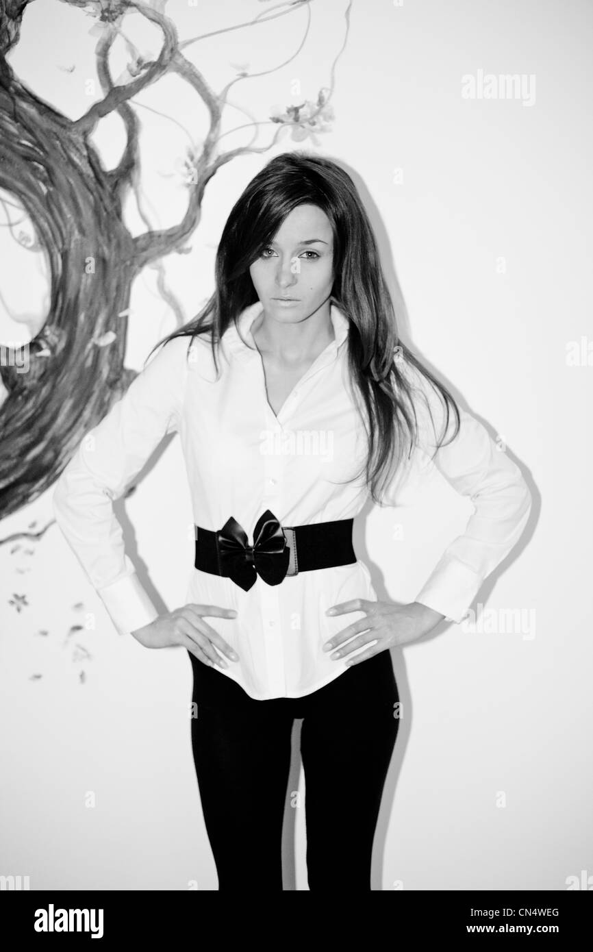 Ein einfarbiges Porträt einer besinnlichen Frau in einer stilvollen weißen Bluse mit schwarzem Gürtel, die vor der künstlerischen Kulisse eines bemalten Baumes steht. Stockfoto