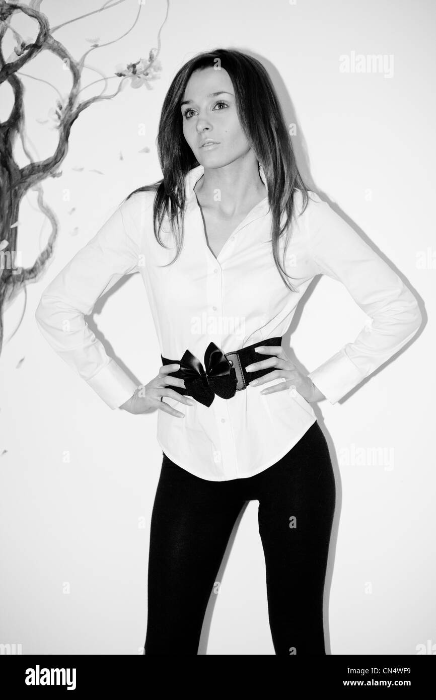 Ein einfarbiges Porträt einer besinnlichen Frau in einer stilvollen weißen Bluse mit schwarzem Gürtel, die vor der künstlerischen Kulisse eines bemalten Baumes steht. Stockfoto