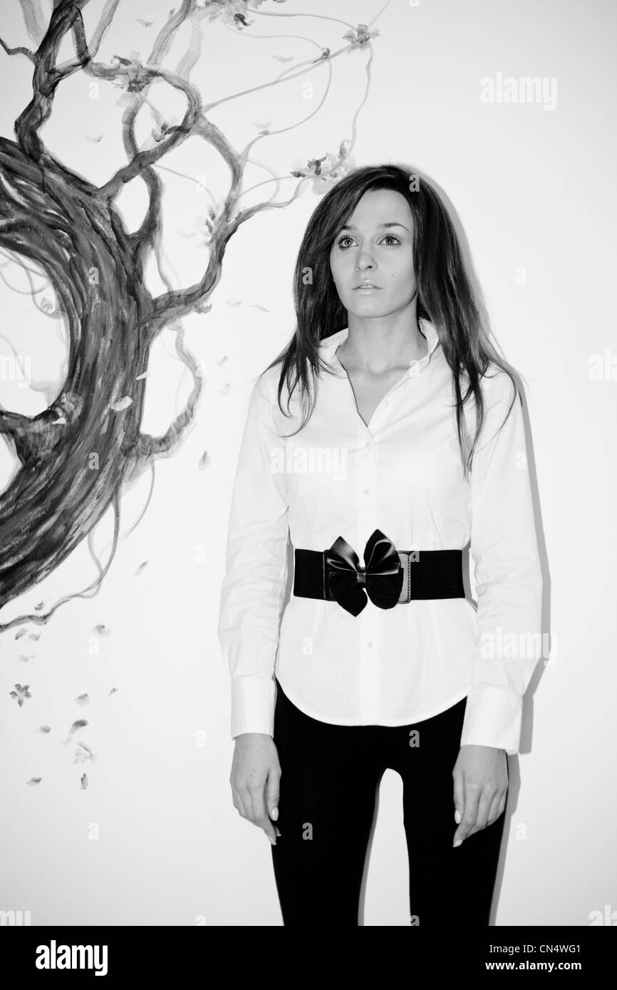 Ein einfarbiges Porträt einer besinnlichen Frau in einer stilvollen weißen Bluse mit schwarzem Gürtel, die vor der künstlerischen Kulisse eines bemalten Baumes steht. Stockfoto